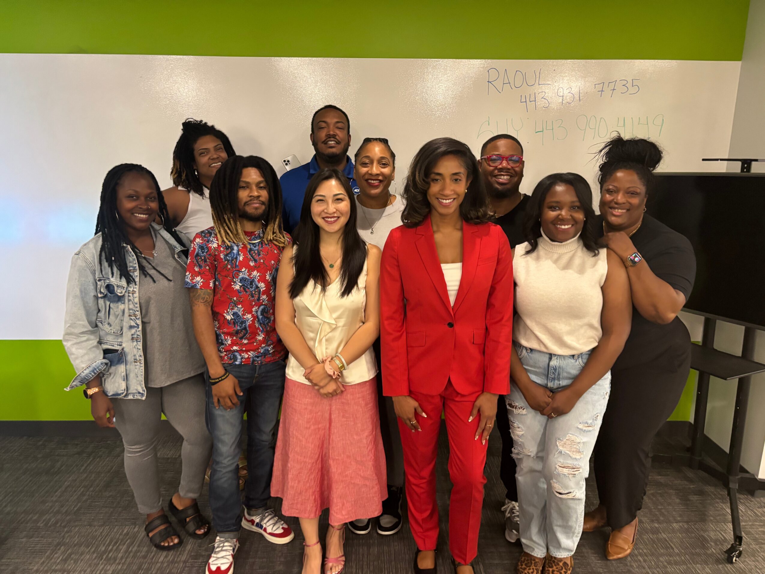 Group of ten diverse people standing together in a room with green and white walls, smiling at the camera.