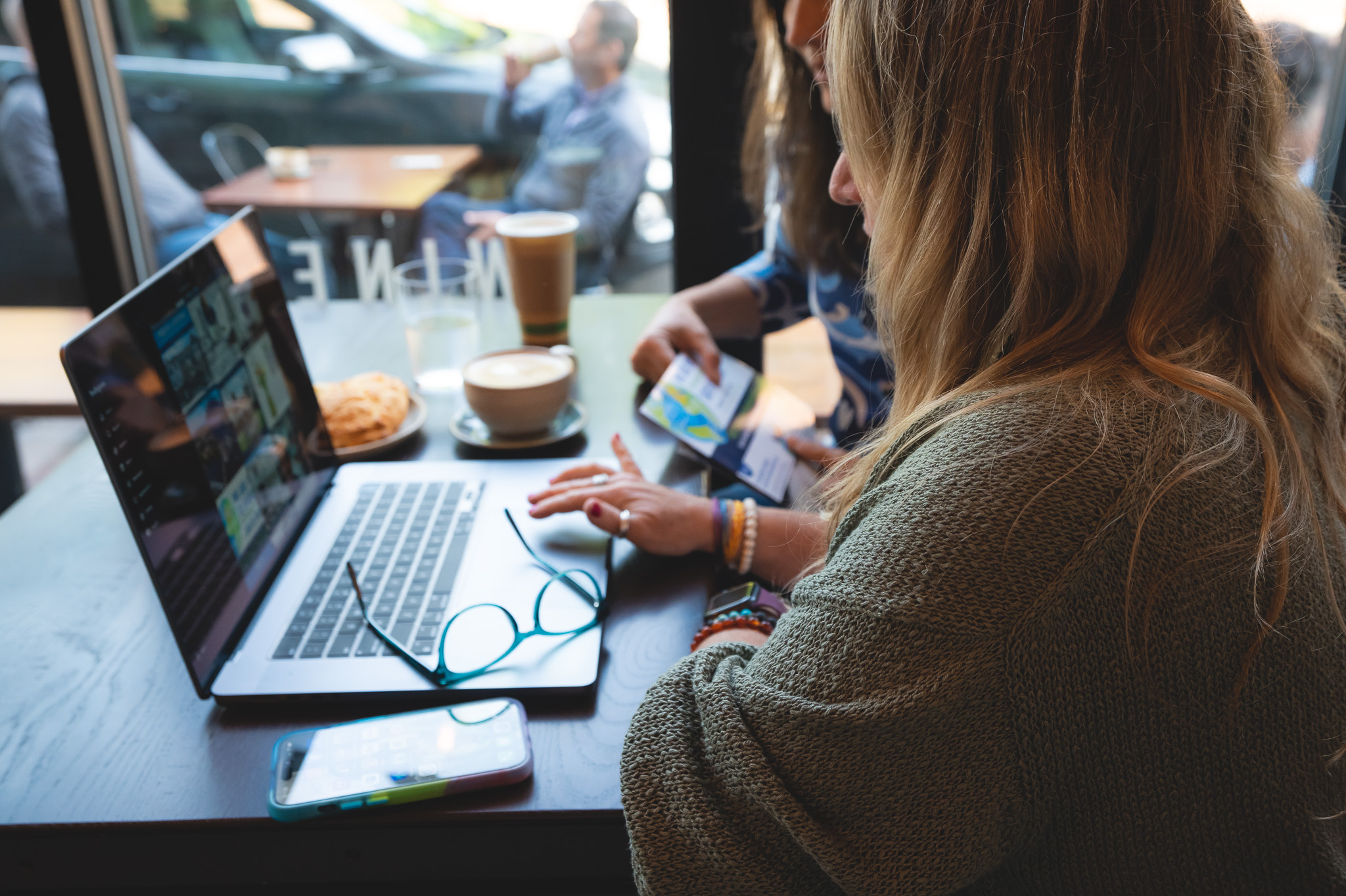 Woman with brown hair working on a laptop at a cafe table, with a person outside visible through the window.