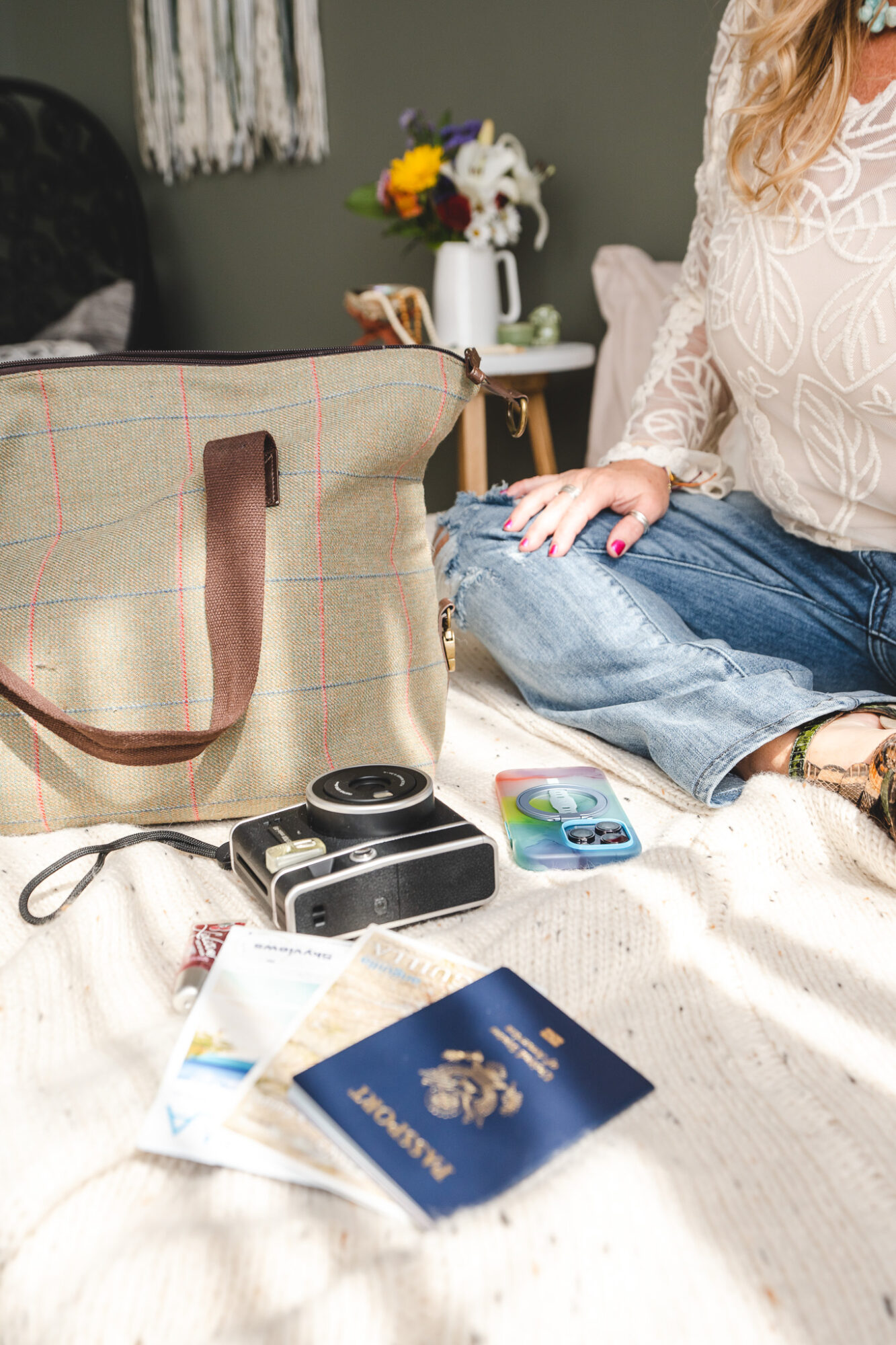 Person sitting on bed with a beige bag, camera, passport, and phone nearby, in a cozy room with flowers in the background.