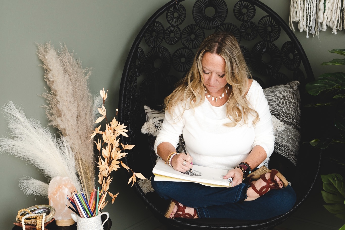 Woman sitting cross-legged on a round black chair, writing in a notebook, with decorative plants and a wall hanging behind her.
