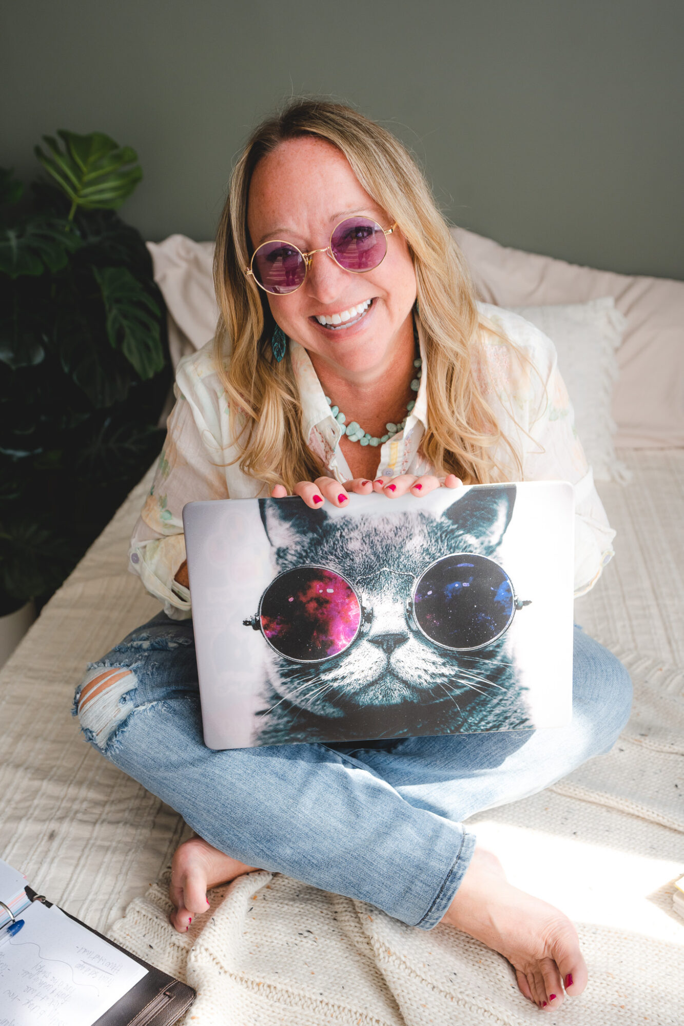 Woman with blonde hair and pink sunglasses sitting on bed with a laptop featuring a cat face design, smiling.