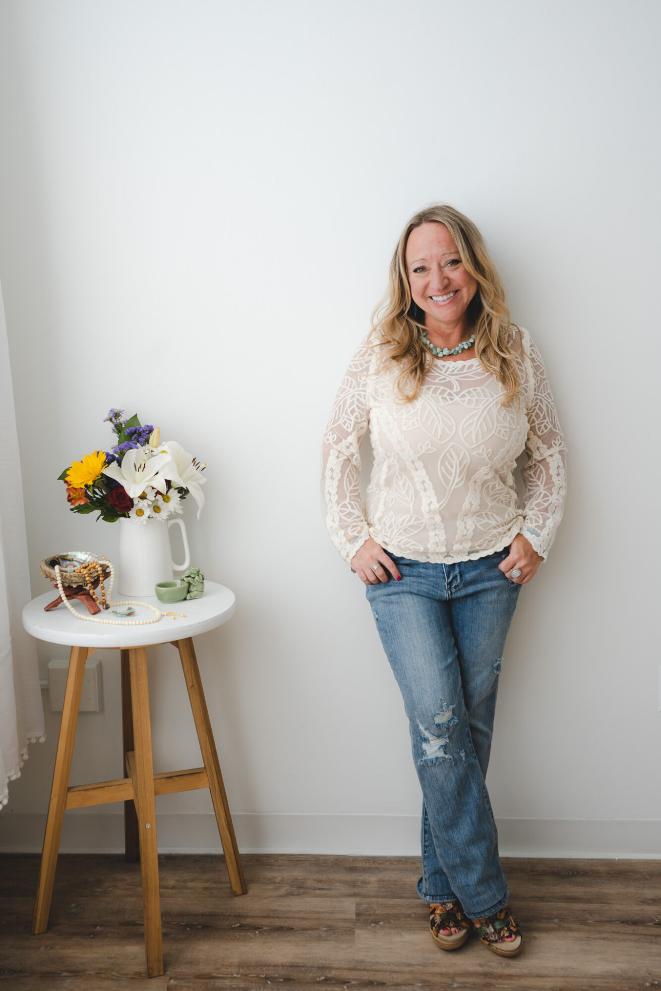Woman with long blonde hair smiling, wearing a white lace top and jeans, standing against a white wall.