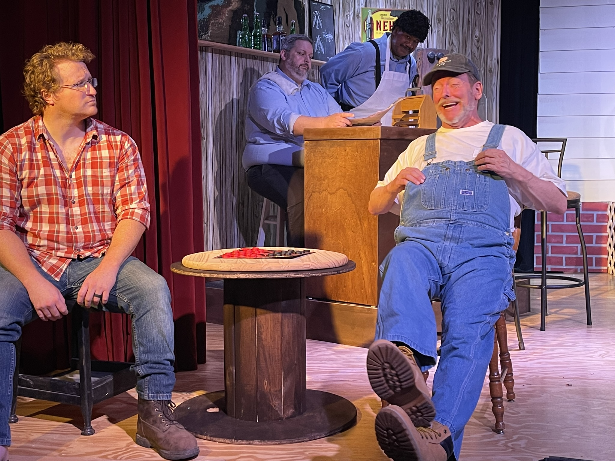 Four people on stage, one sitting, one standing, and two behind a counter, with a small round table in front.
