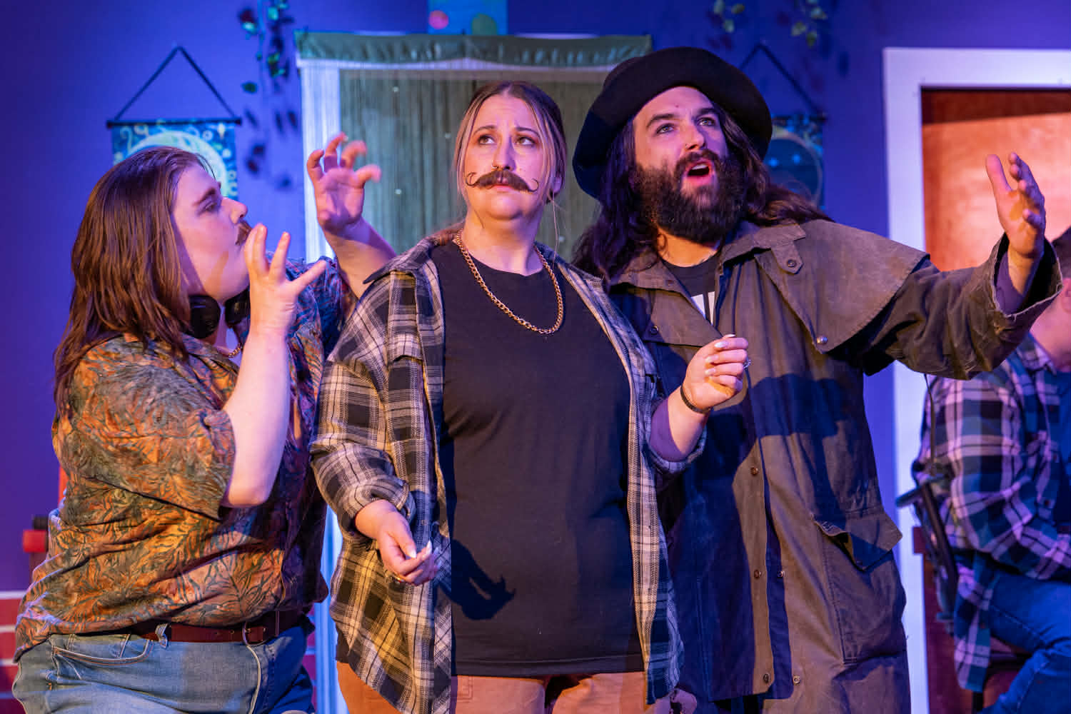 Three people on stage, one with a fake mustache, gesturing and talking, with a colorful background and props.