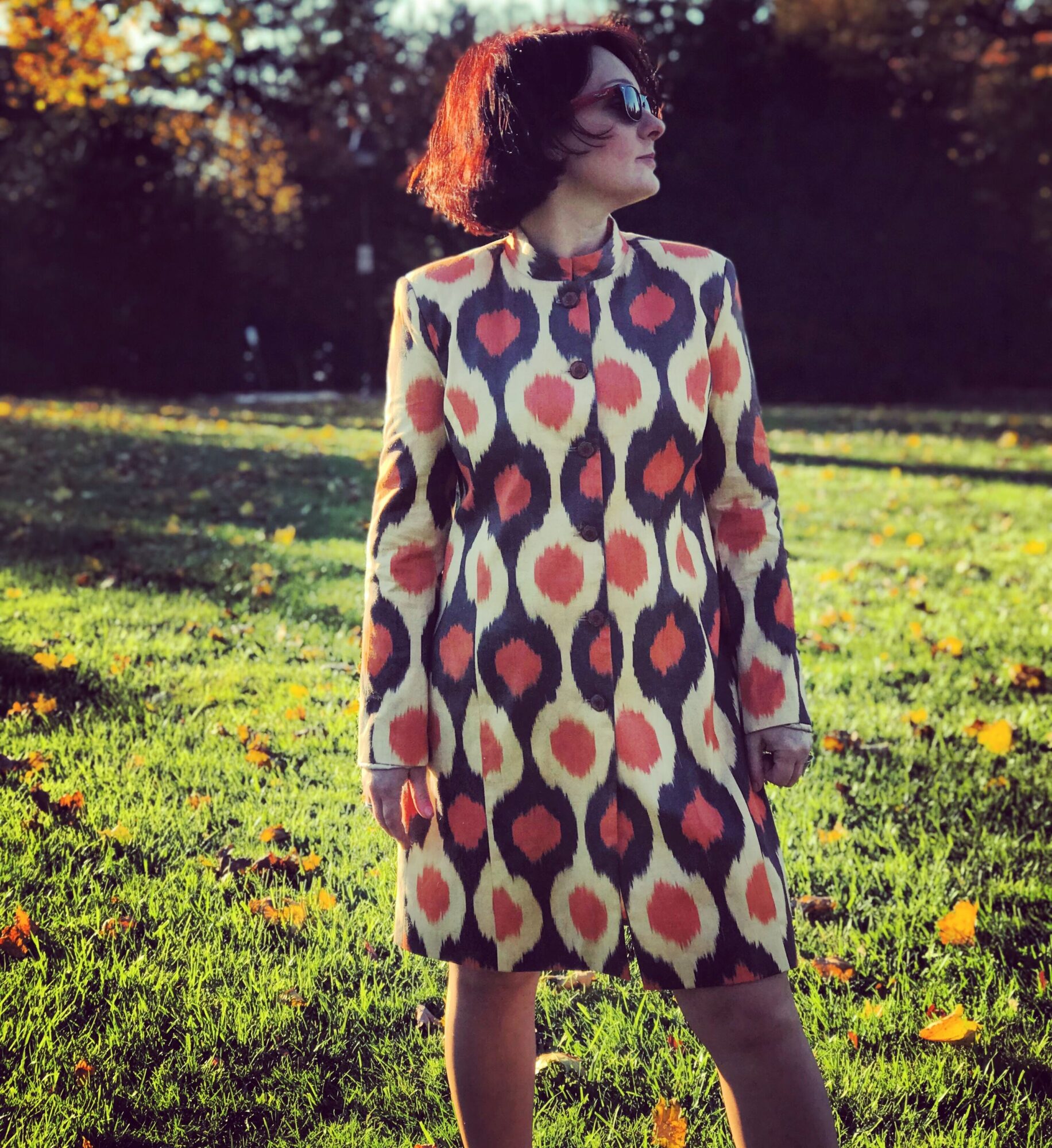 Woman with short hair and sunglasses standing outdoors in a grassy area with trees, wearing a patterned coat.