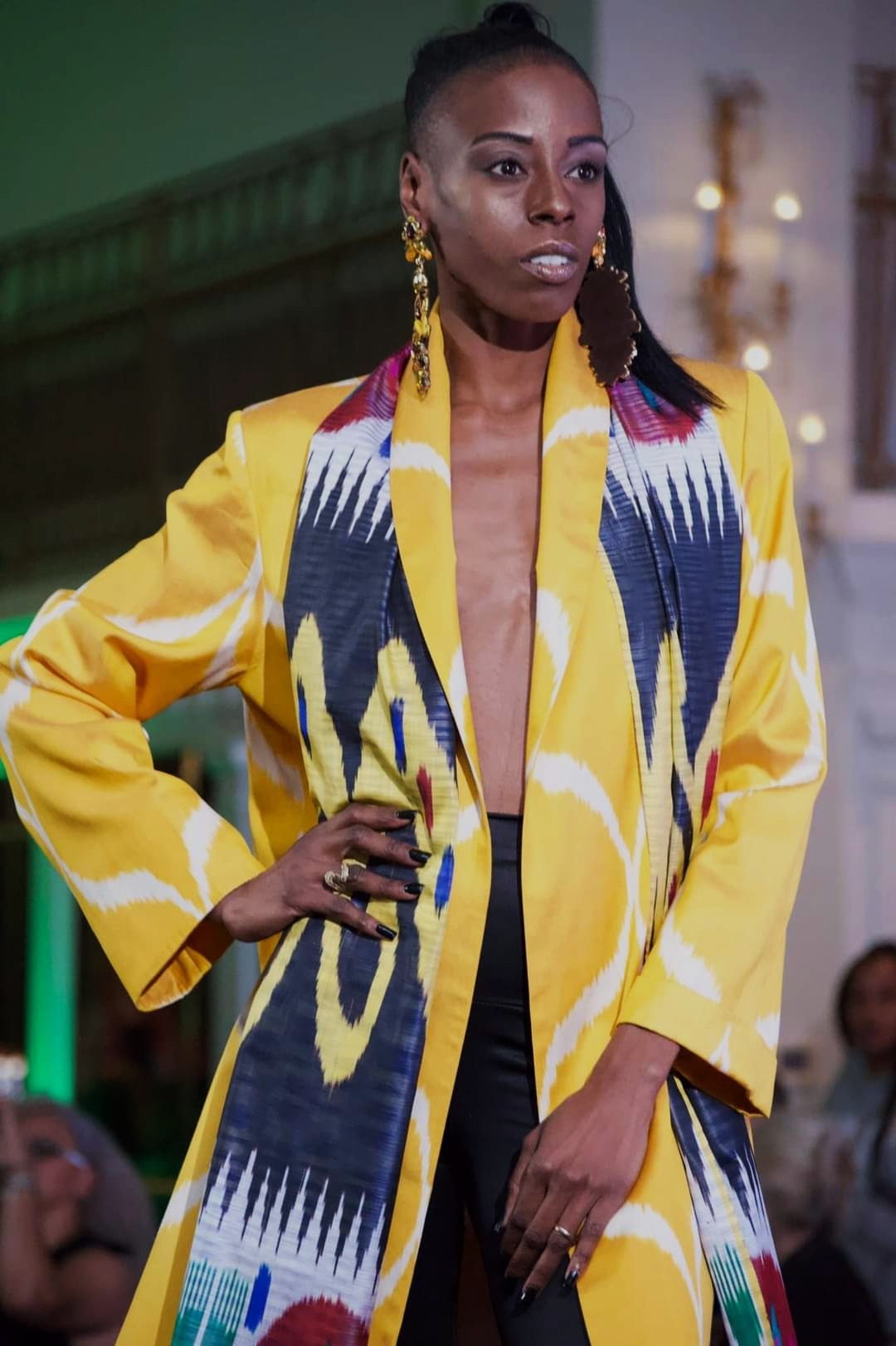 Woman in a yellow and blue patterned coat with hand on hip, standing indoors with blurred background