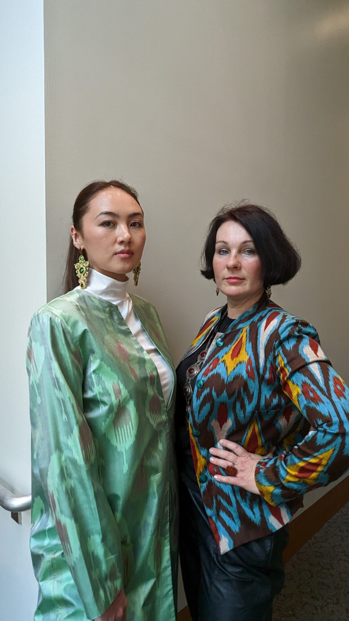 Two women standing side by side, one in a shiny green dress and the other in a colorful patterned top, against a plain background.