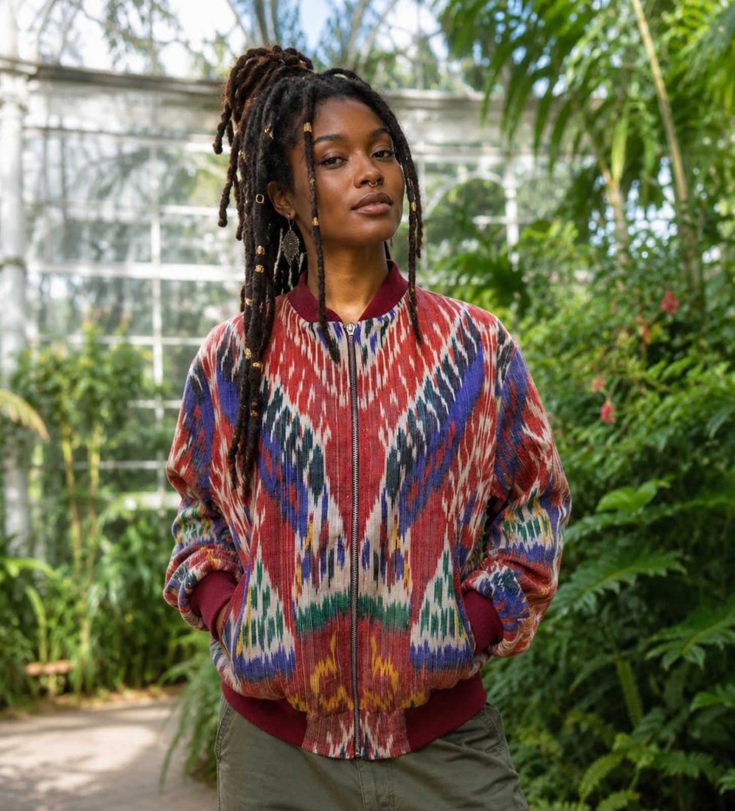 Young woman with dreadlocks in a colorful patterned jacket outdoors surrounded by greenery.
