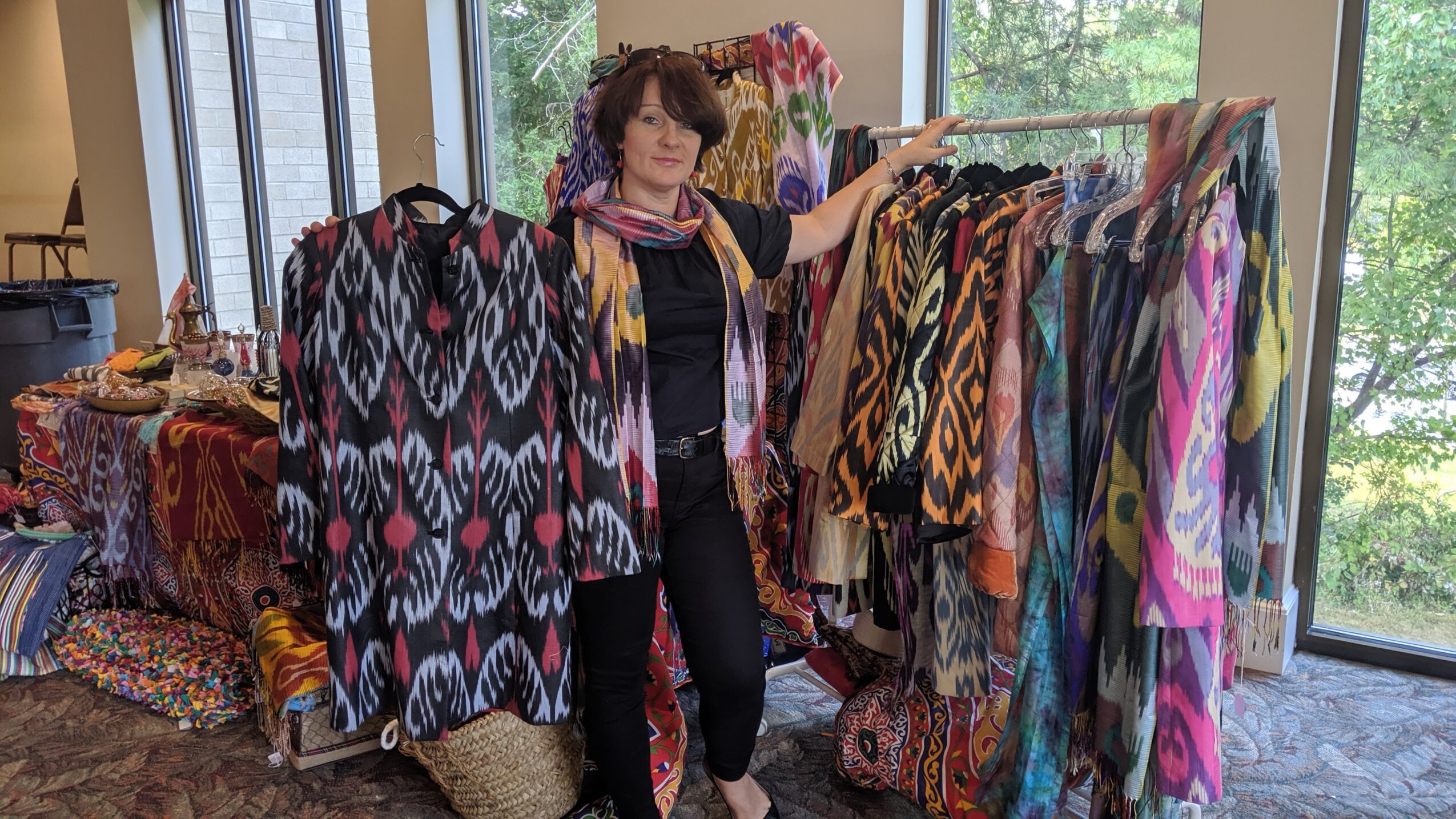 Woman browsing colorful clothing racks indoors near large windows, with trees visible outside.