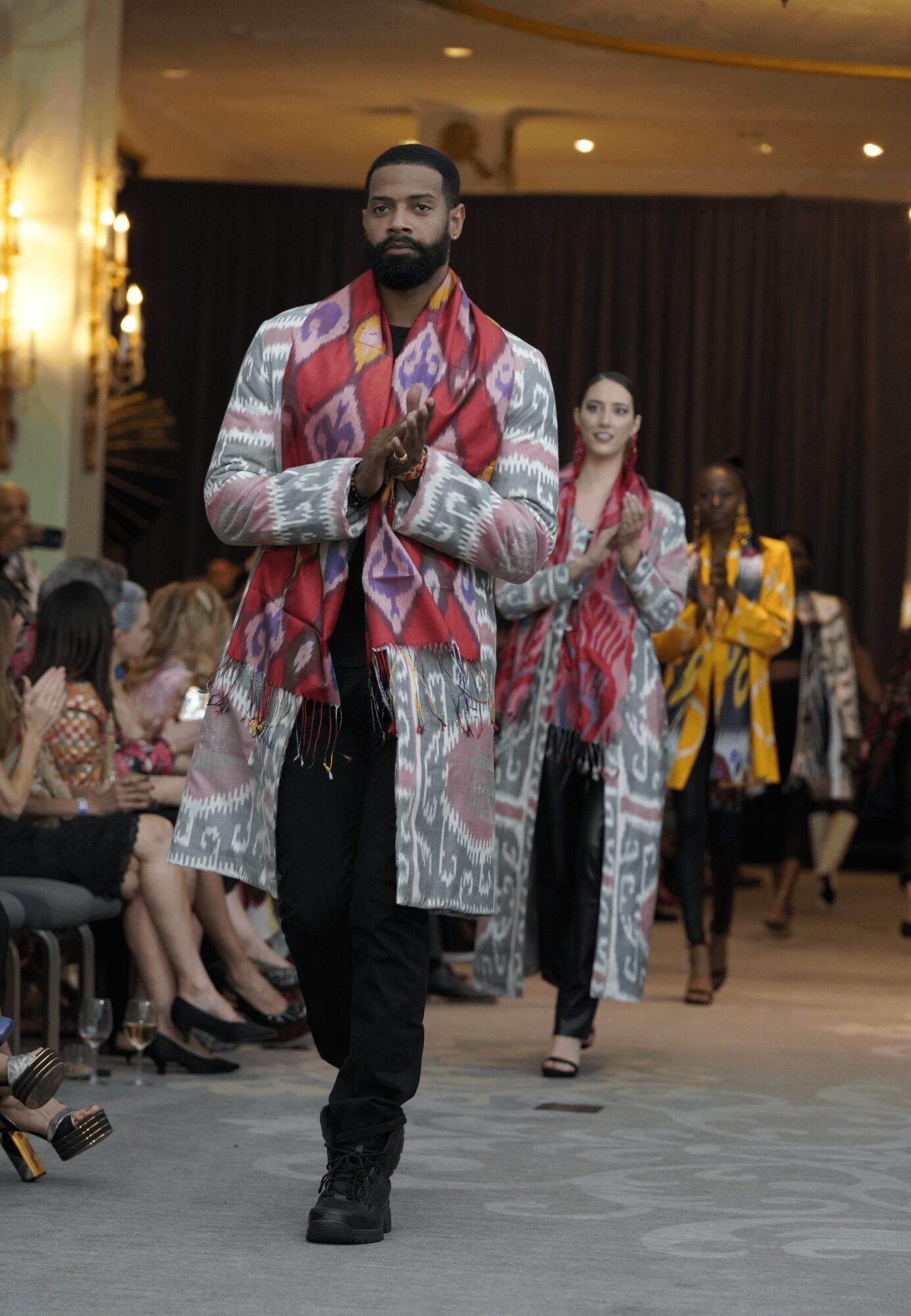 Man walking on runway with hands together, wearing a patterned coat and scarf, audience seated on sides, dim lighting.