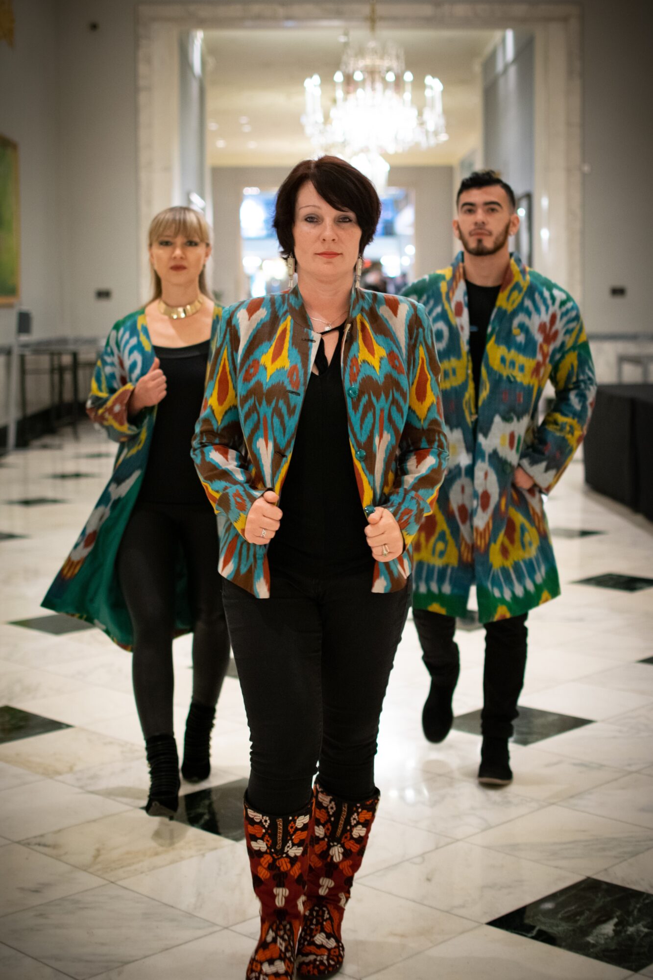 Four people walking in a hotel lobby with a chandelier, wearing colorful patterned coats and black clothing.