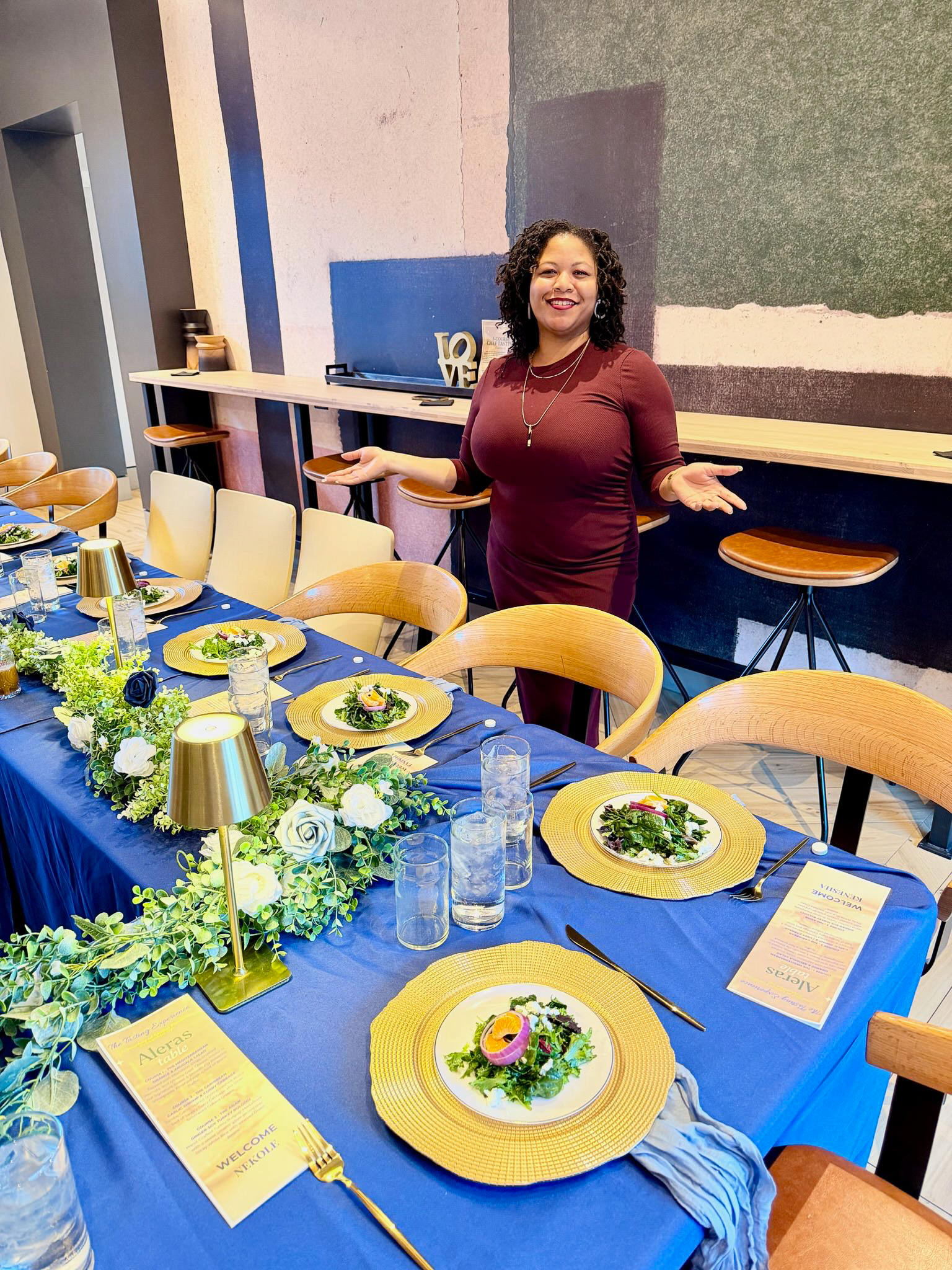 Woman with curly hair in a maroon dress stands behind a decorated dining table, smiling with arms outstretched, in a room with blue and green walls.
