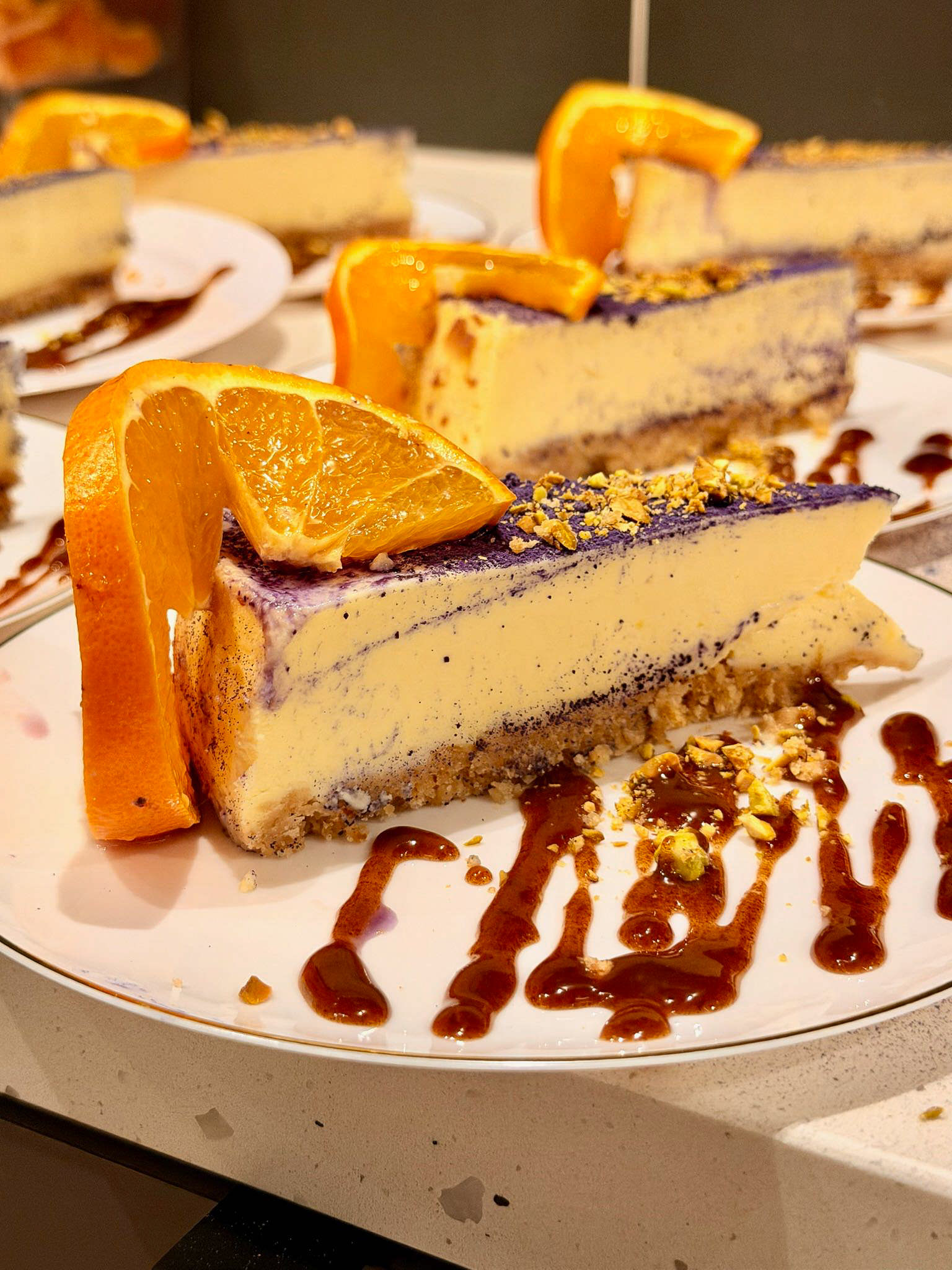 Slices of cheesecake topped with orange slices and garnished with chocolate sauce and crushed nuts on a white plate.