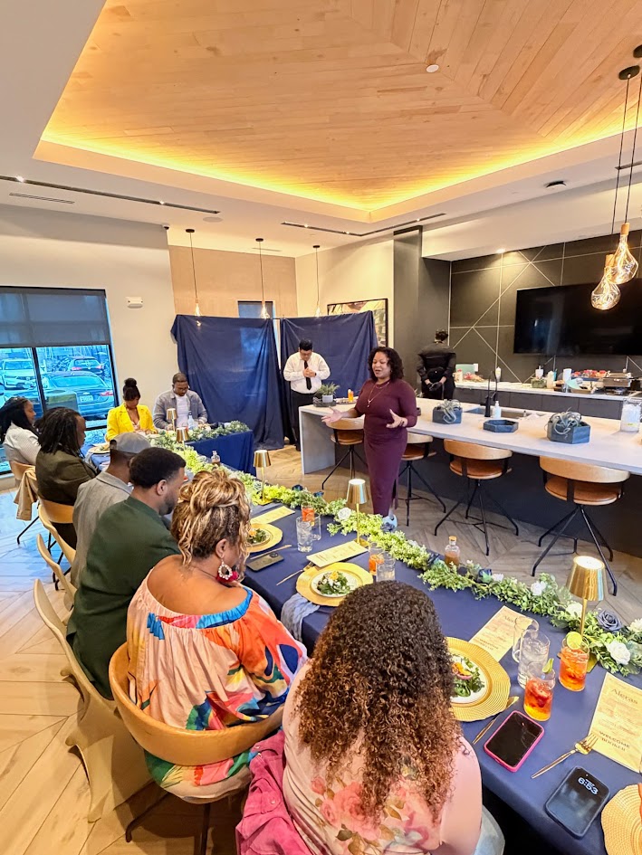 People seated at a long table in a modern restaurant, listening to a woman speaking, with a bar area in the background.