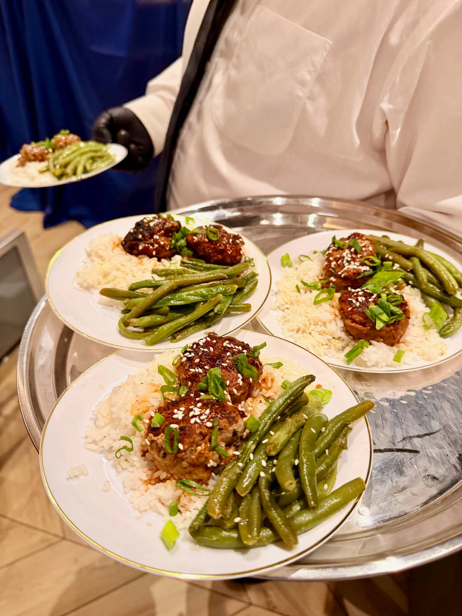 Three plates of food with green beans, rice, and meat sauce on a tray, held by a person in a white shirt.