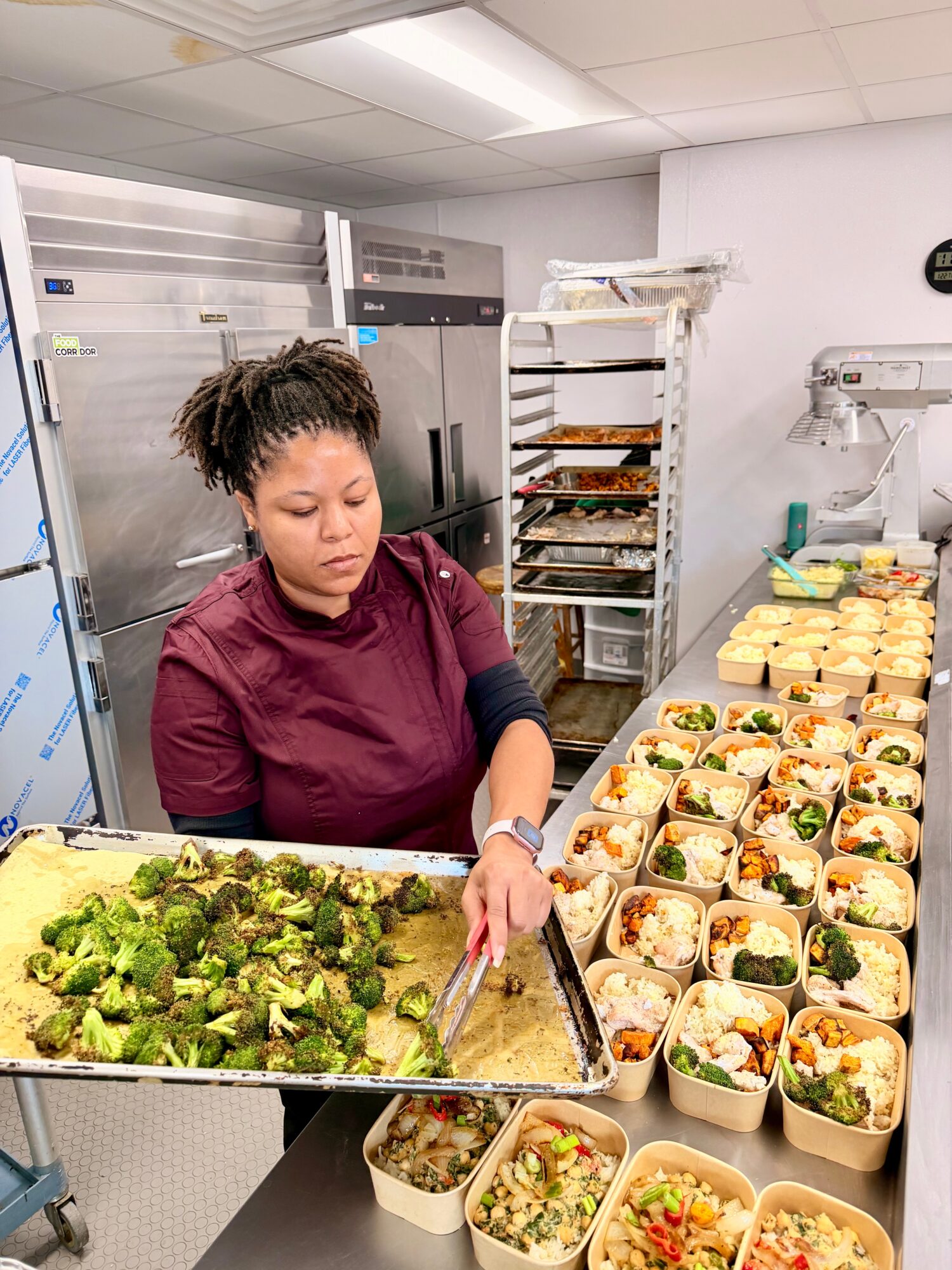Woman in maroon uniform prepares food in a kitchen with trays of salads and sandwiches on a table.