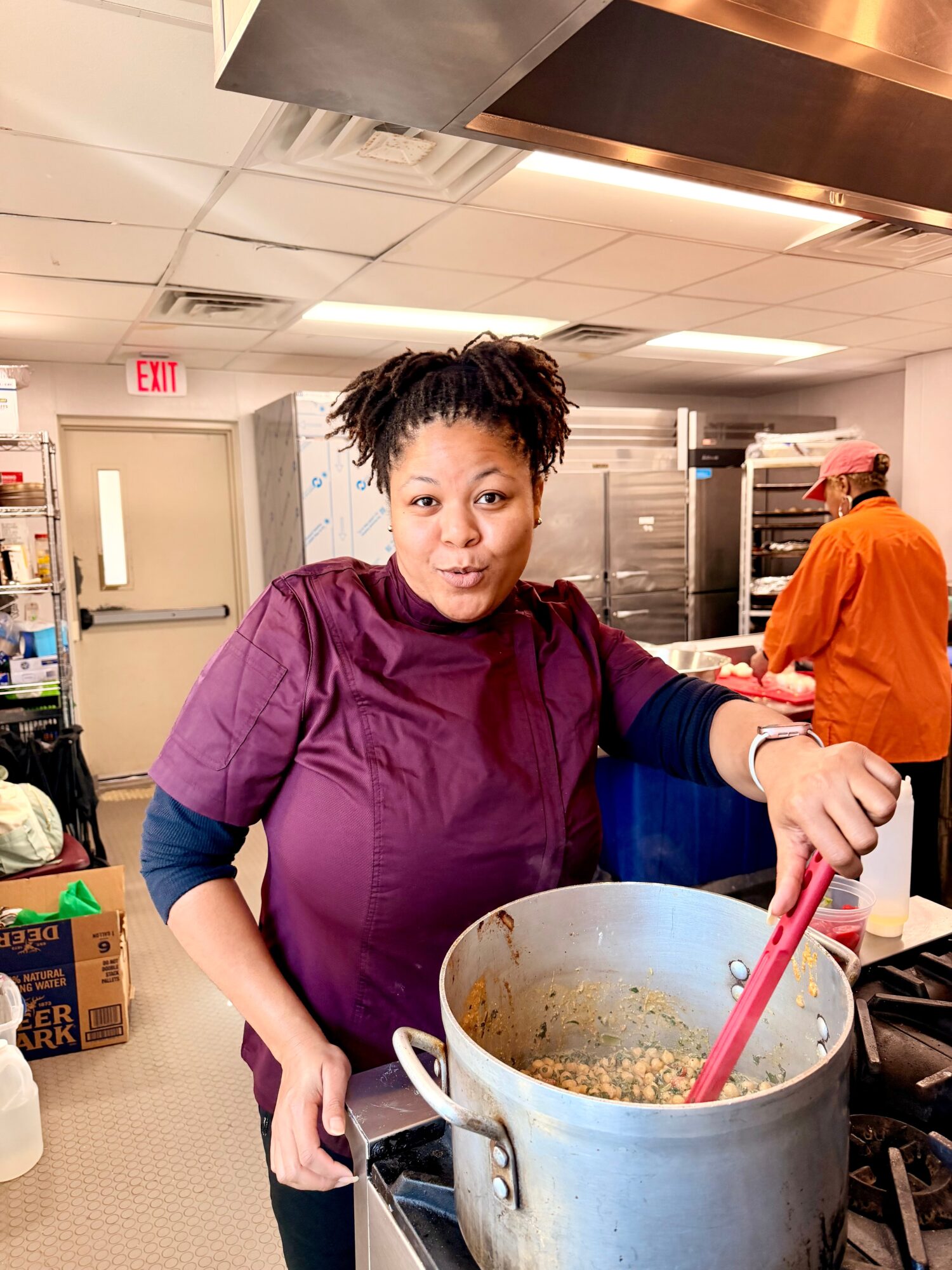 Woman in purple scrubs stirring a large pot in a kitchen, with another person in orange in the background.