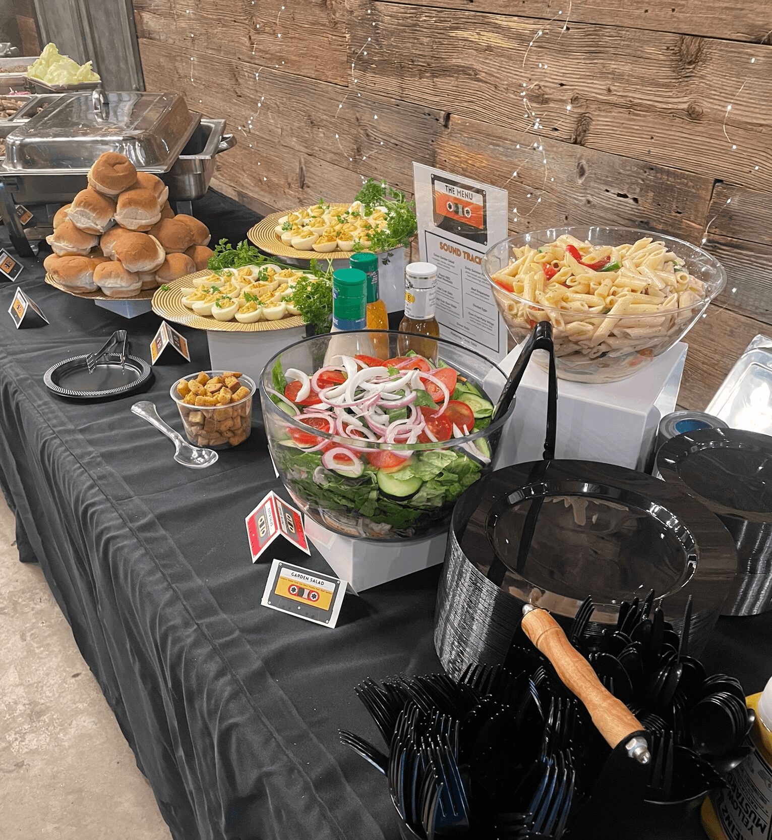 Buffet table with salads, bread rolls, and condiments against a wooden wall, with utensils and labels visible.