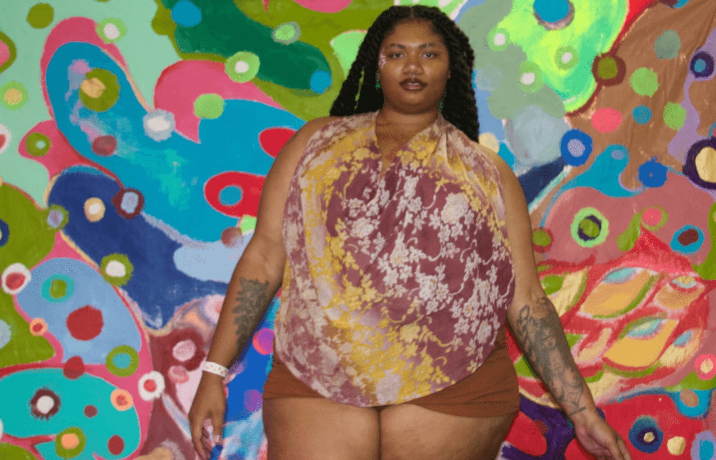 Person with long dark hair and tattoos on arms standing in front of a colorful abstract background.