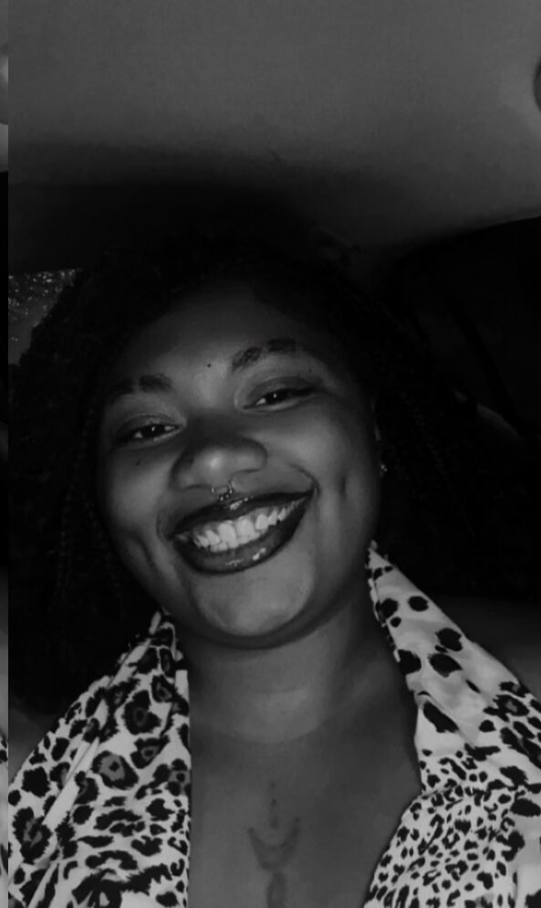 Smiling person with curly hair, wearing a patterned shirt, in a car interior, in black and white.
