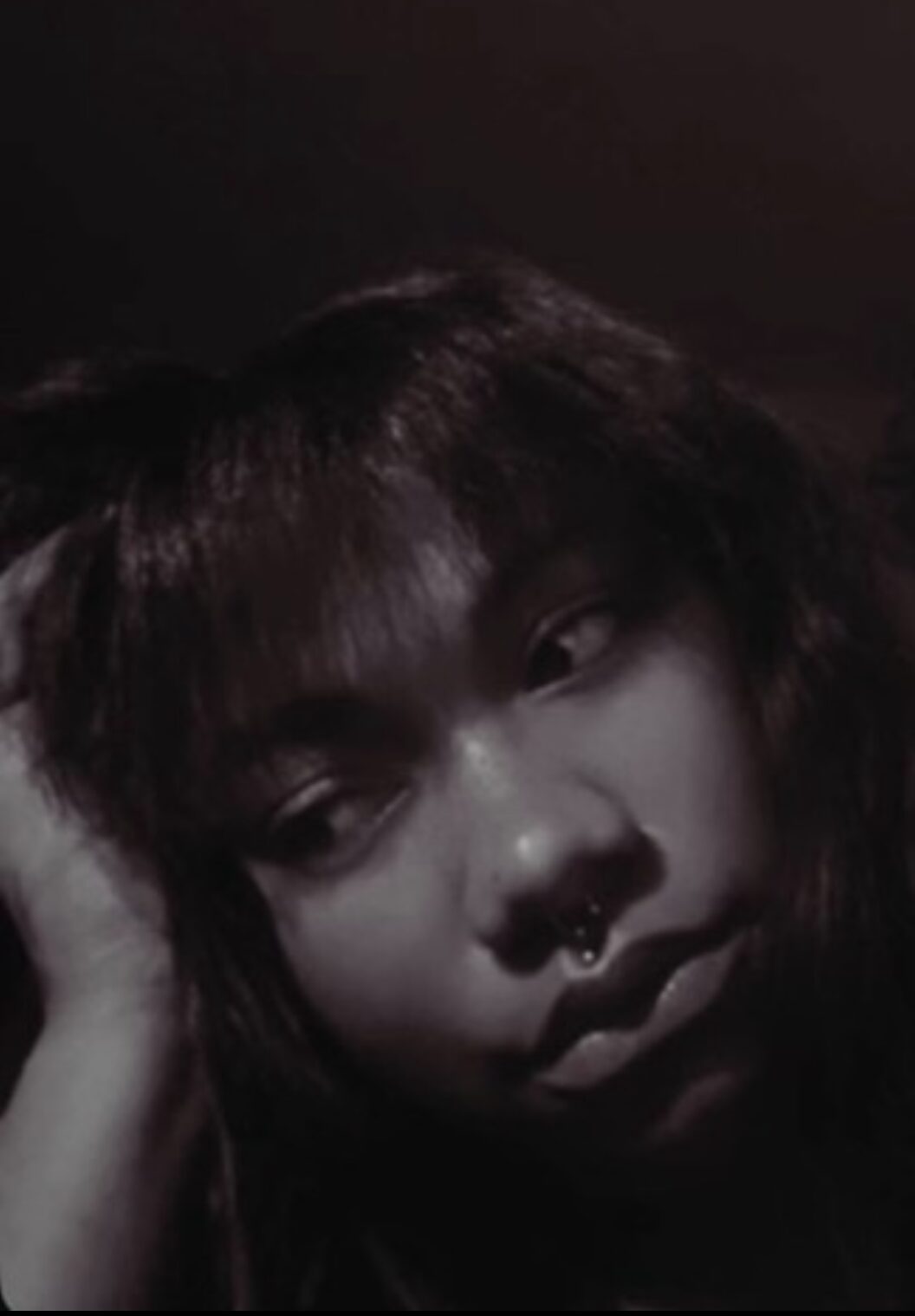 Close-up of a person with dark hair and a septum piercing, resting their head on their hand, in black and white.