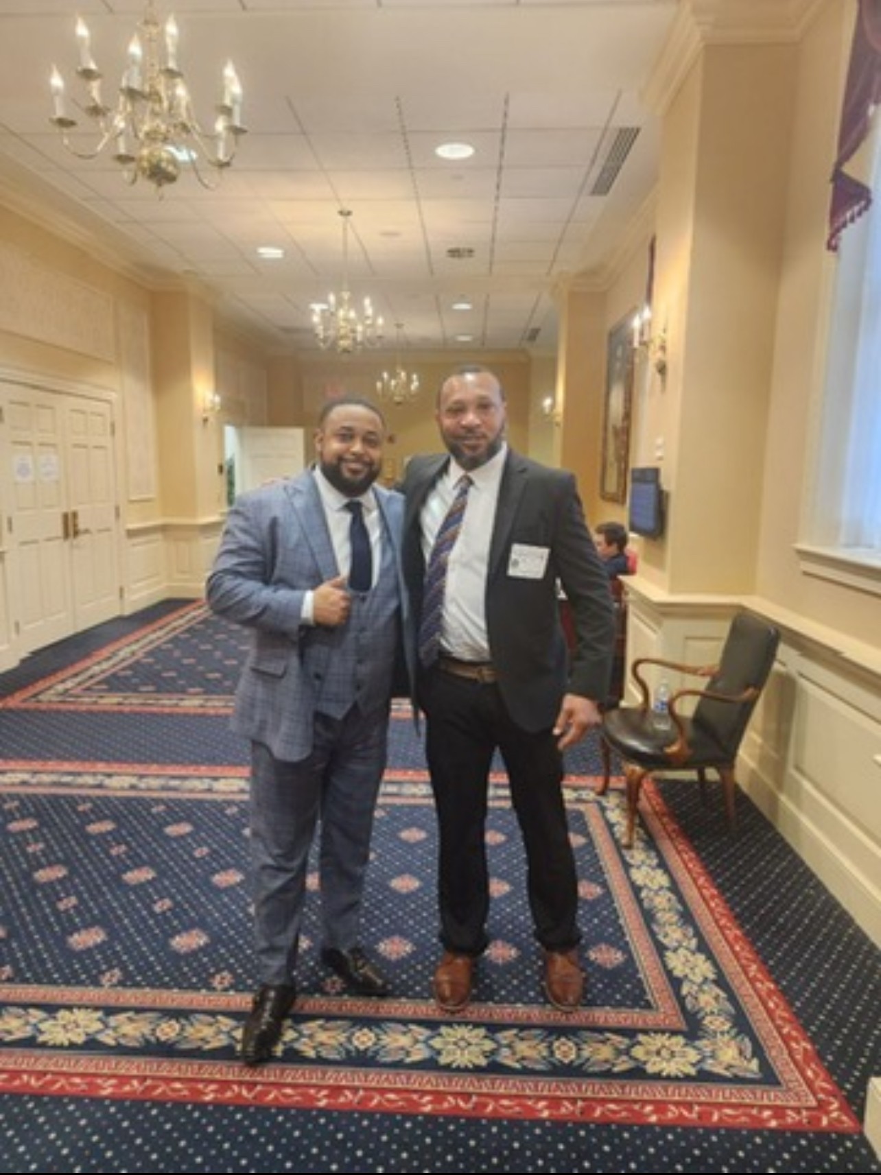 Two men in suits standing in a decorated hallway with chandeliers and a patterned carpet.