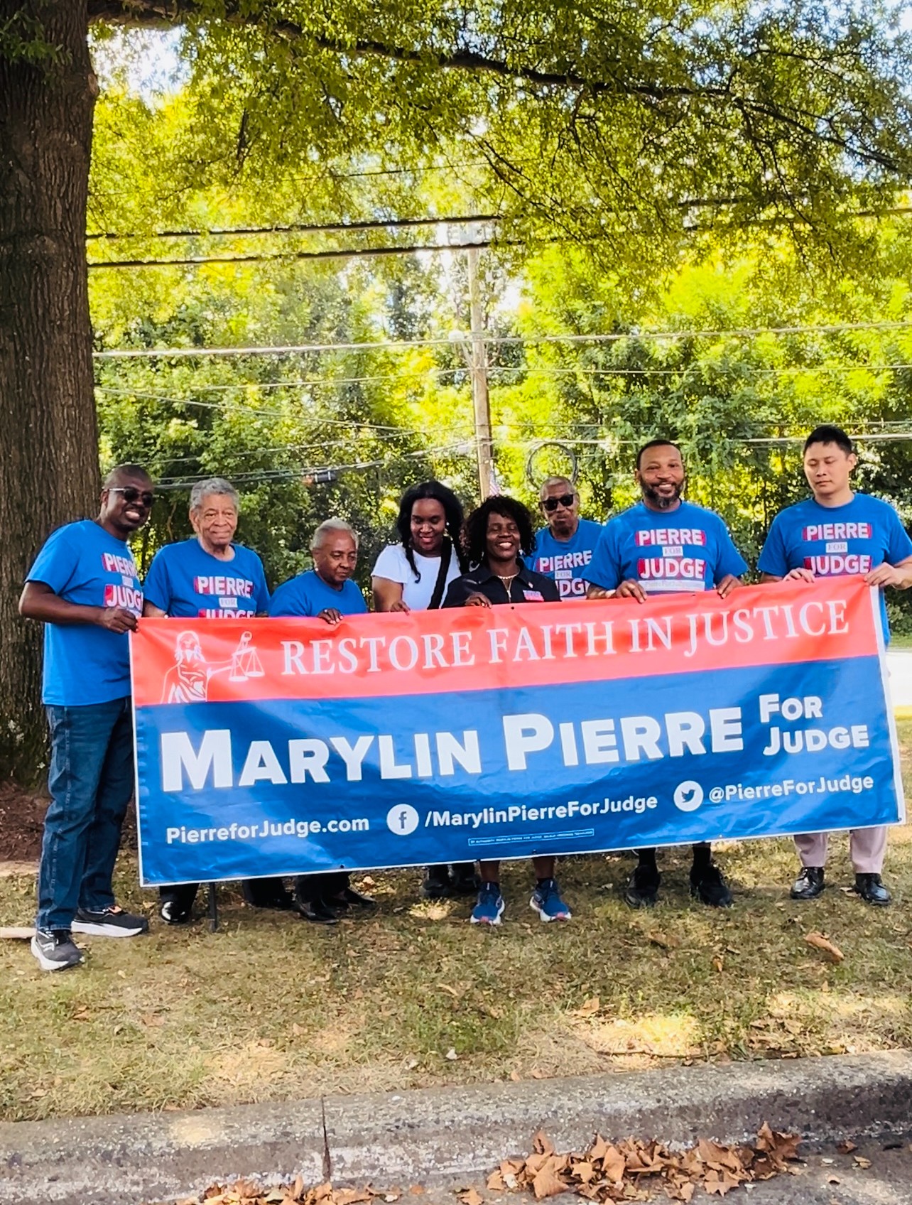 Group of people holding a blue and red banner outdoors under trees, supporting Marylin Pierre for judge.