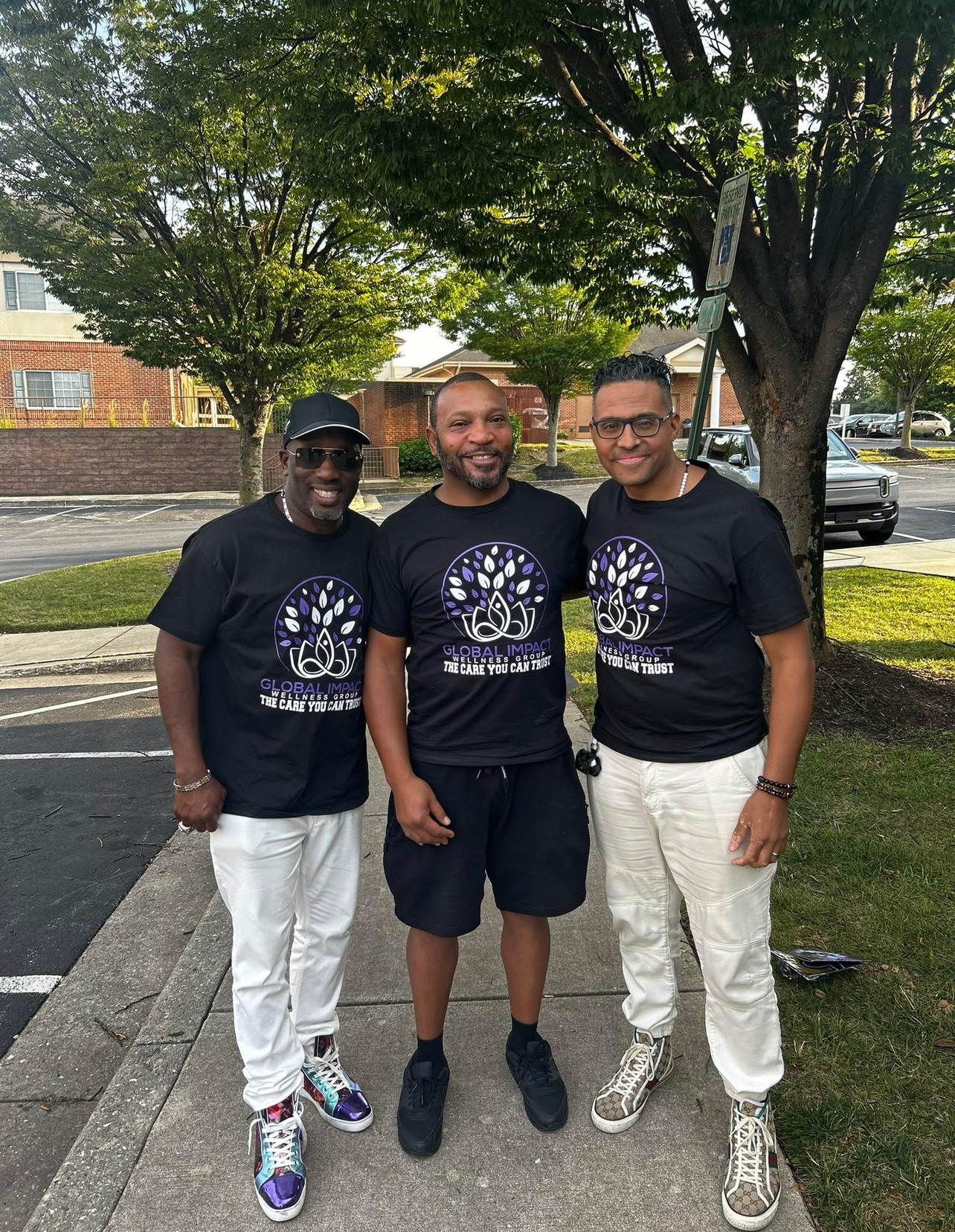 Three men standing outdoors under a tree, smiling, wearing matching black T-shirts with a circular logo, and casual pants or shorts.