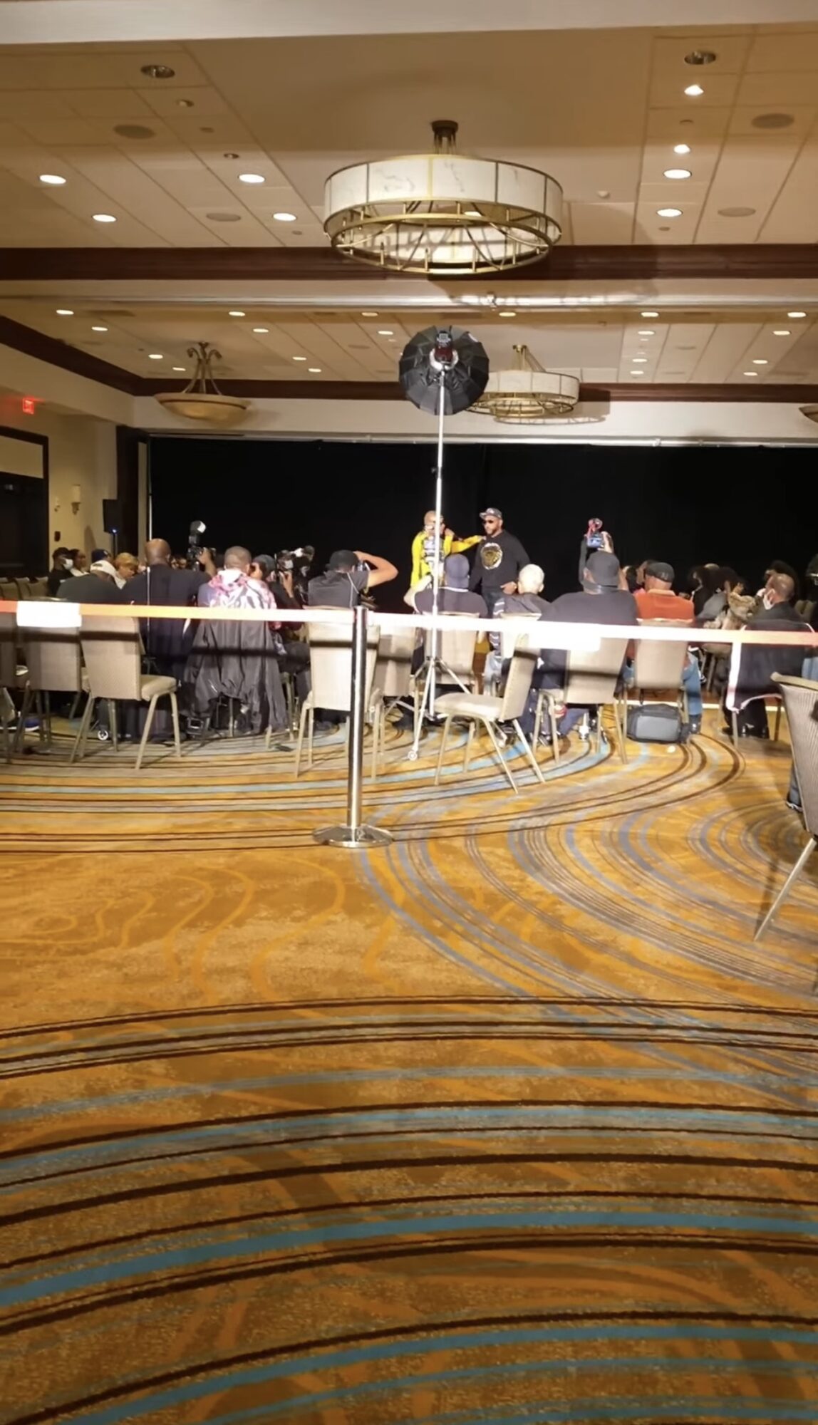 People seated at tables in a large banquet hall with a chandelier, some standing and talking, with a stage in the background.
