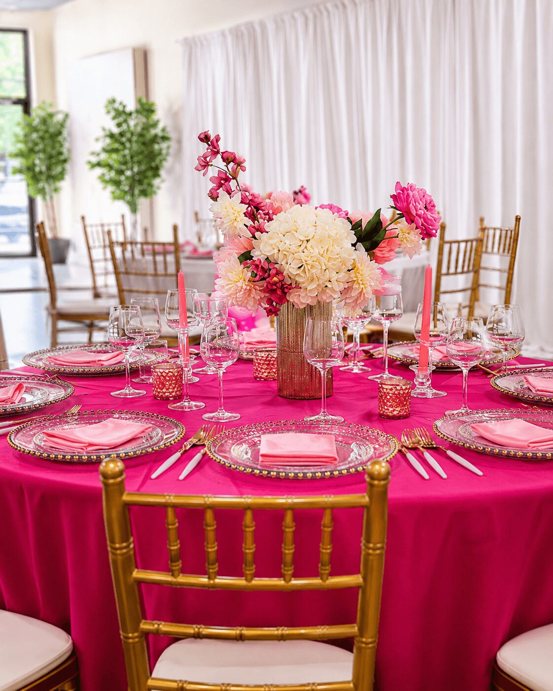 Round table with pink tablecloth, floral centerpiece, pink candles, and elegant place settings in a decorated room.
