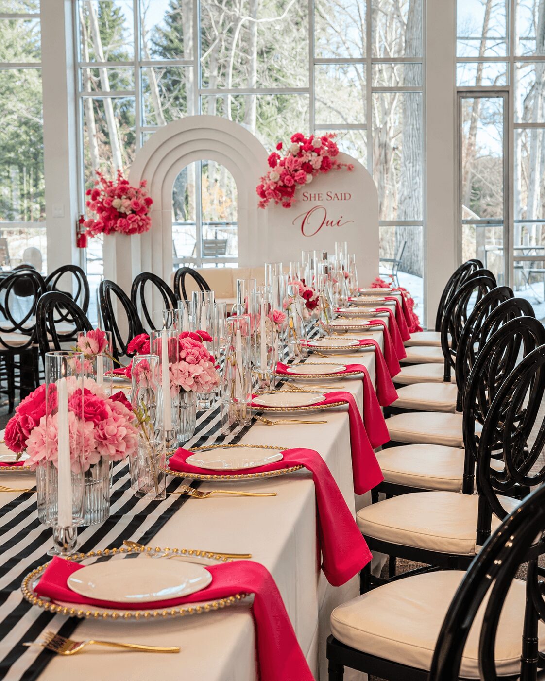 Elegant banquet table set with pink and white flowers, gold cutlery, and pink napkins, in a bright room with large windows.