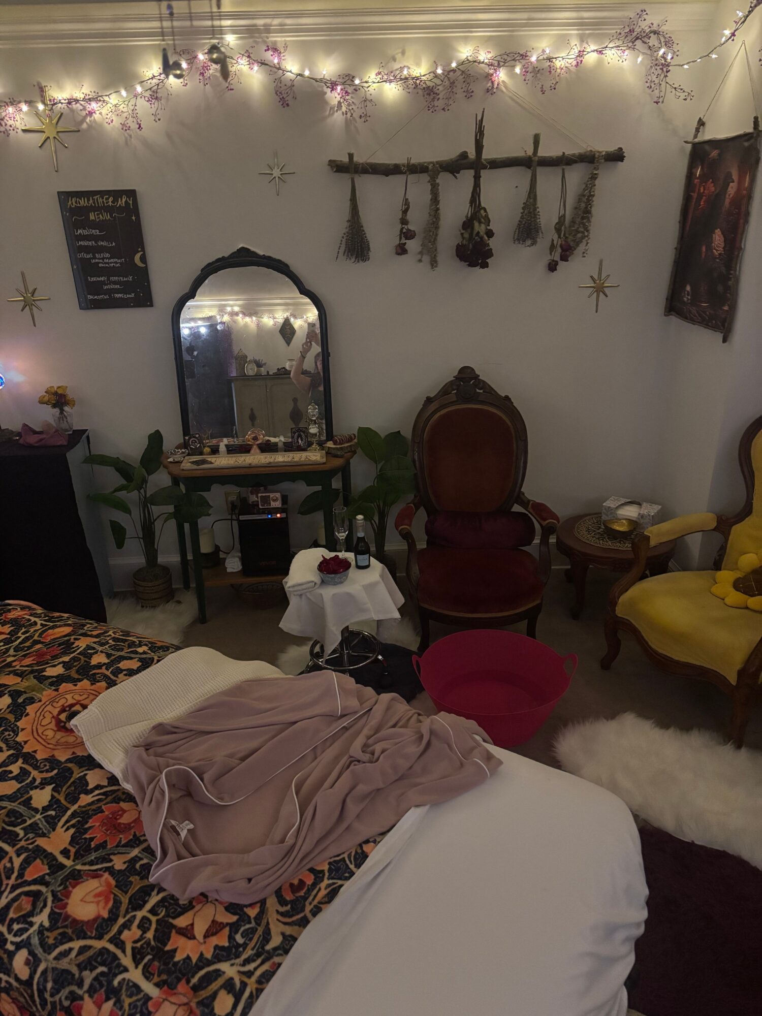 Cozy room with a mirror, chairs, and decorative wall hanging, seen from a bed with clothes and patterned blanket.