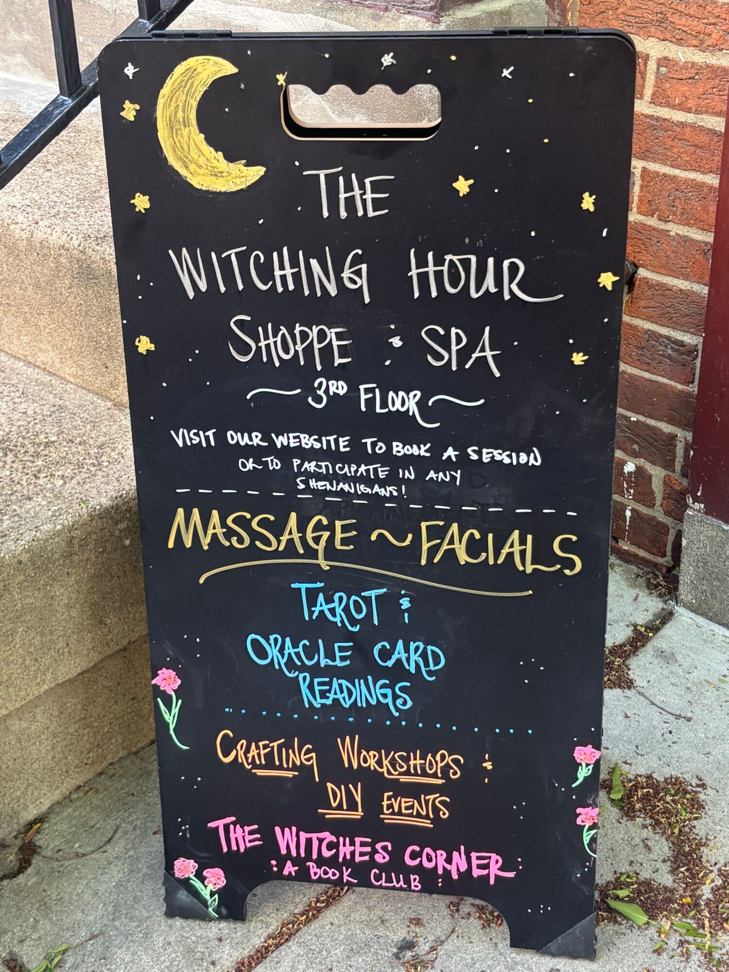 Black signboard with colorful handwritten text and illustrations, including a moon and stars, advertising a witching hour spa and massage services.