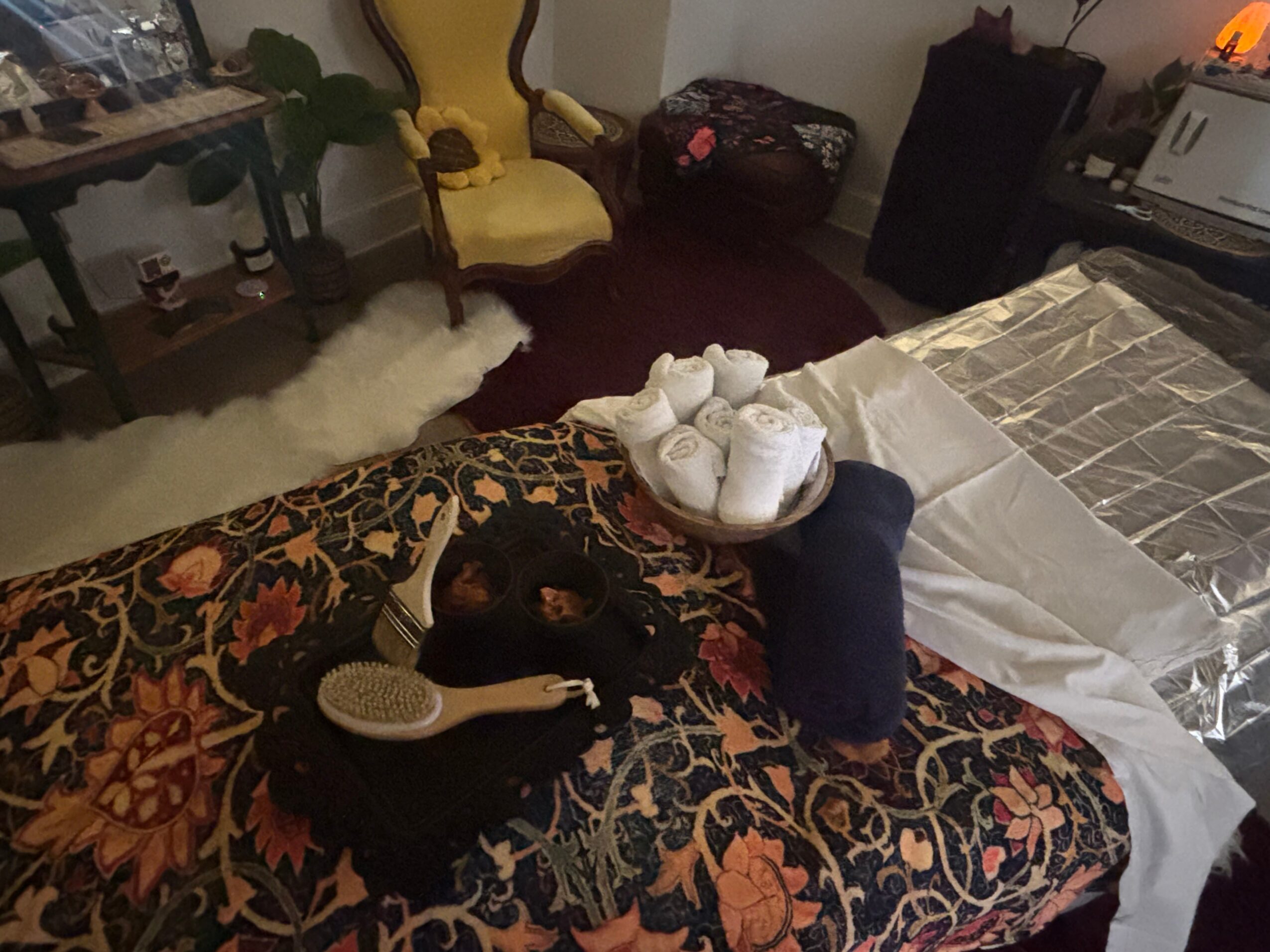 A bed with a floral blanket, a black cat, a hairbrush, and a rolled towel with slippers on top, in a cozy room.