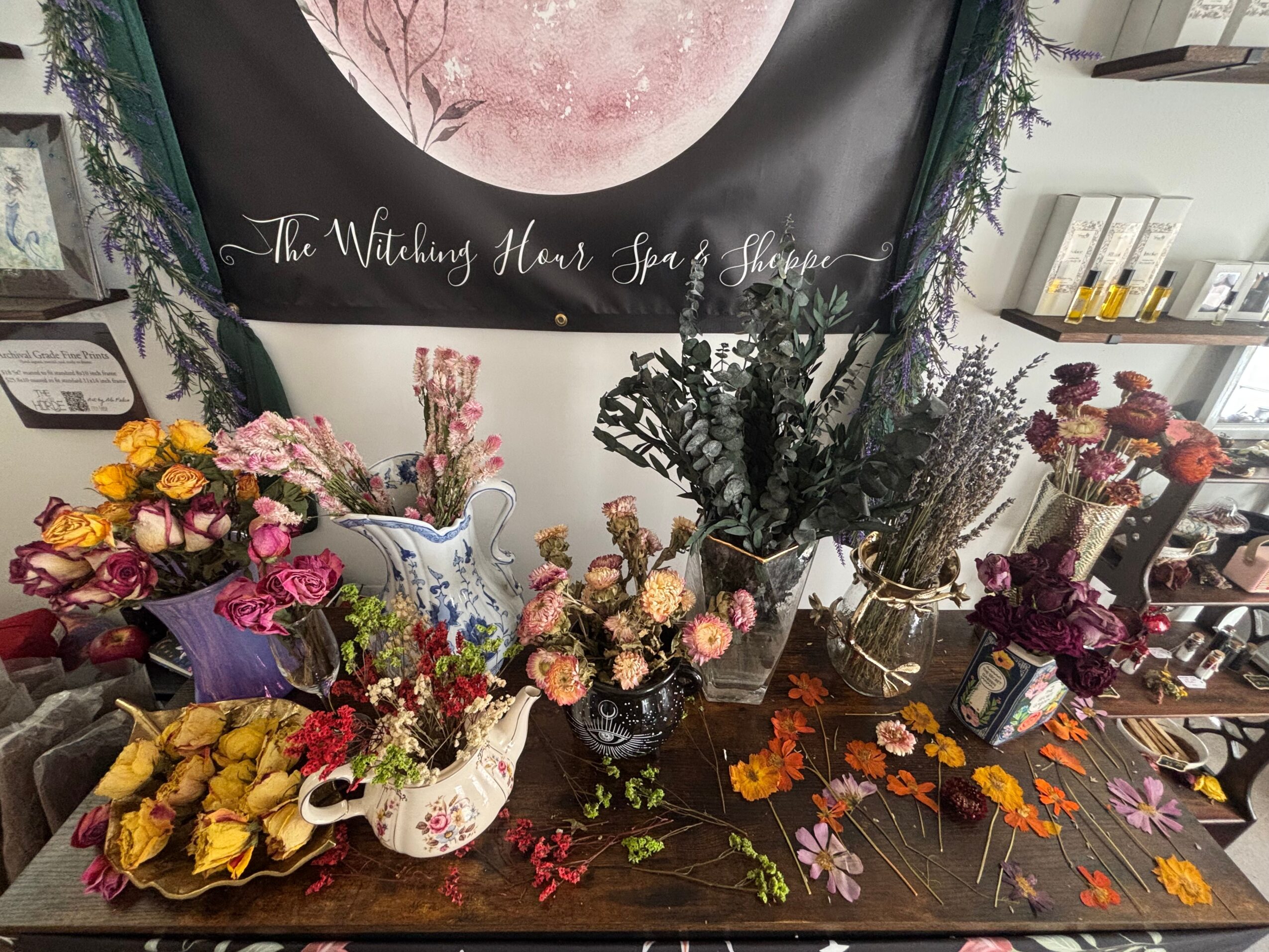 Table with various colorful flower arrangements in vases and containers, with a black banner featuring a pink moon and text in the background.