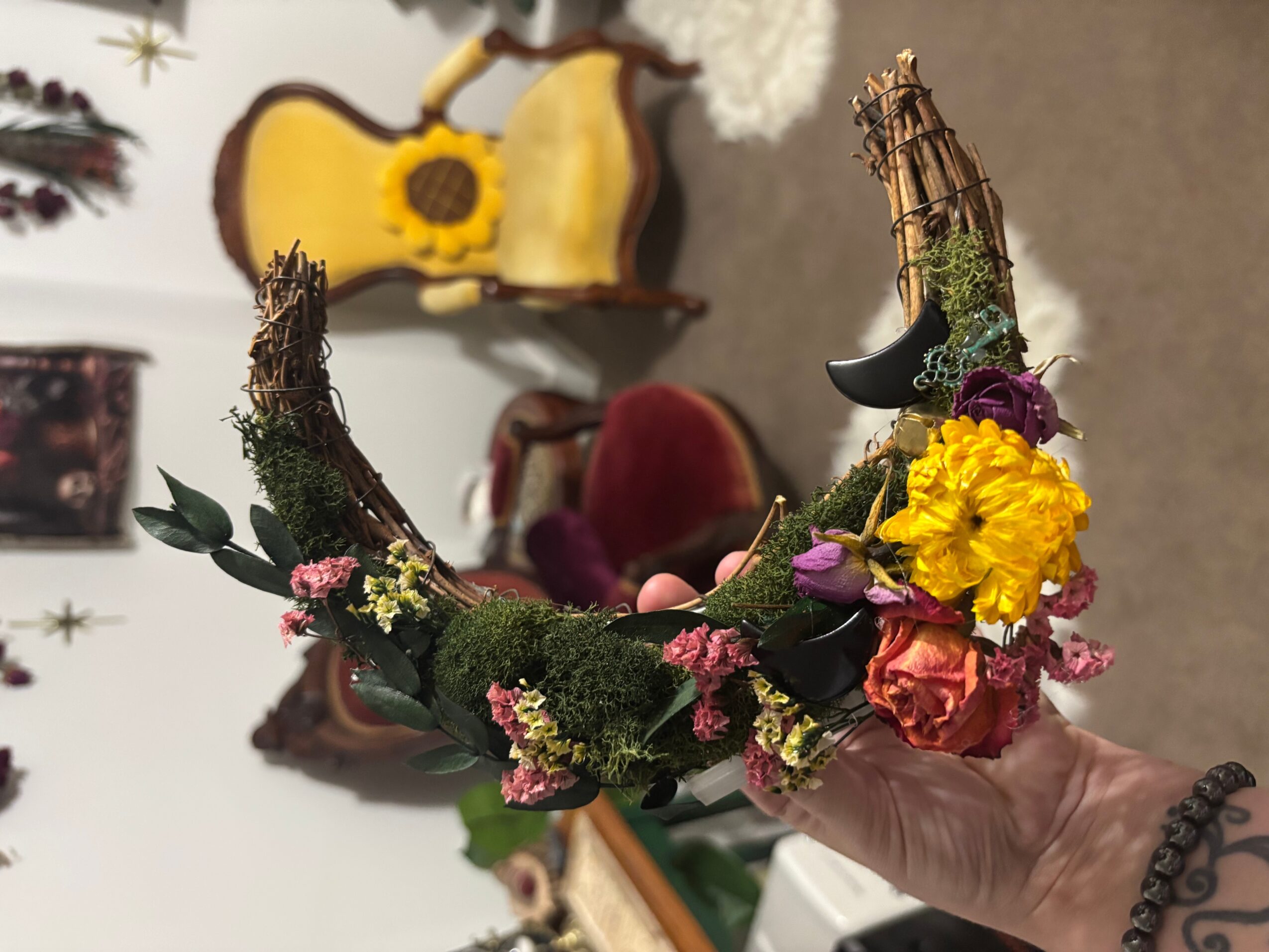 Hand holding a floral crescent-shaped arrangement with moss, flowers, and twigs, in a room with furniture and a guitar in the background.