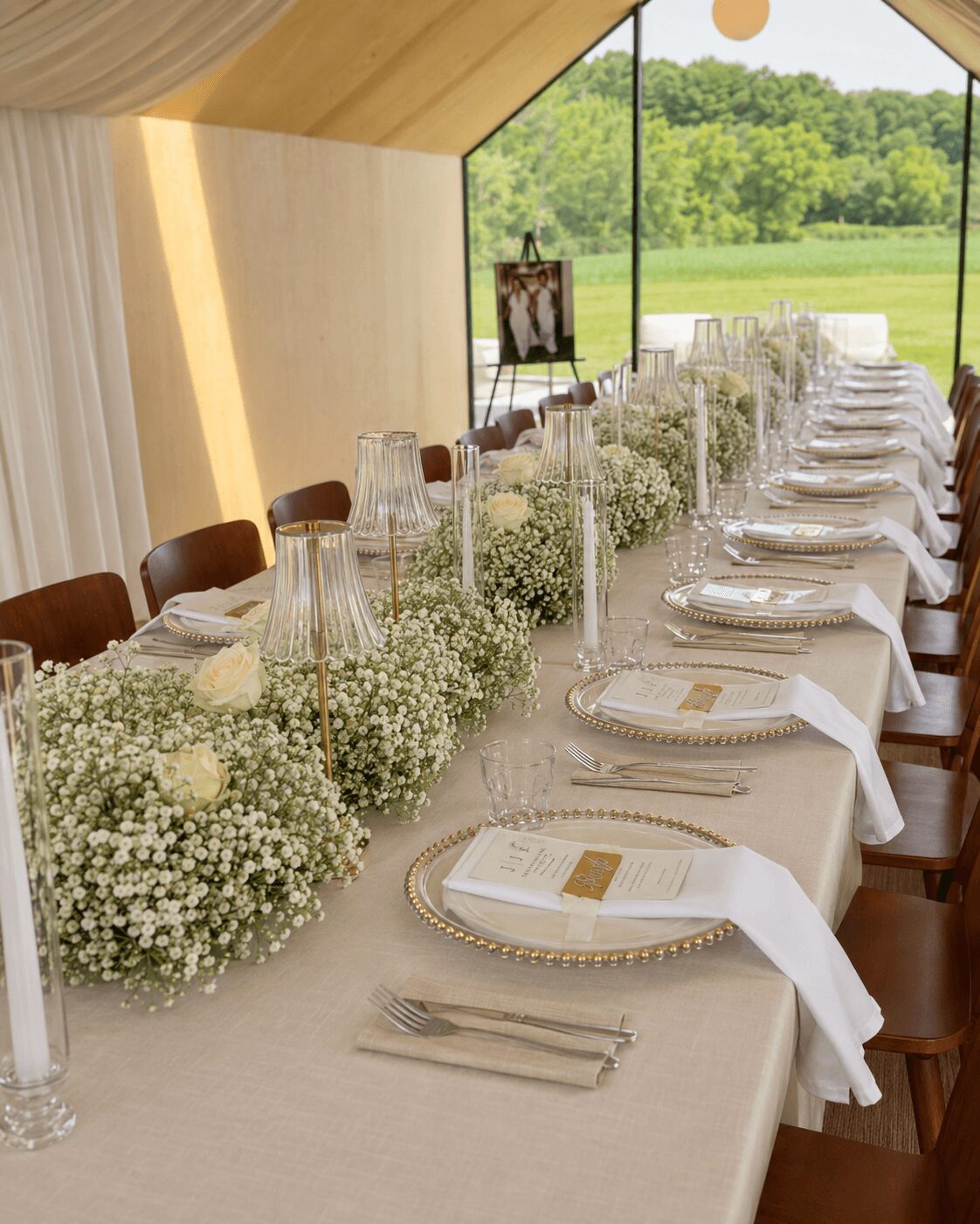 Long dining table with white tablecloth, floral centerpiece, glassware, plates, and napkins, set outdoors with green landscape view.