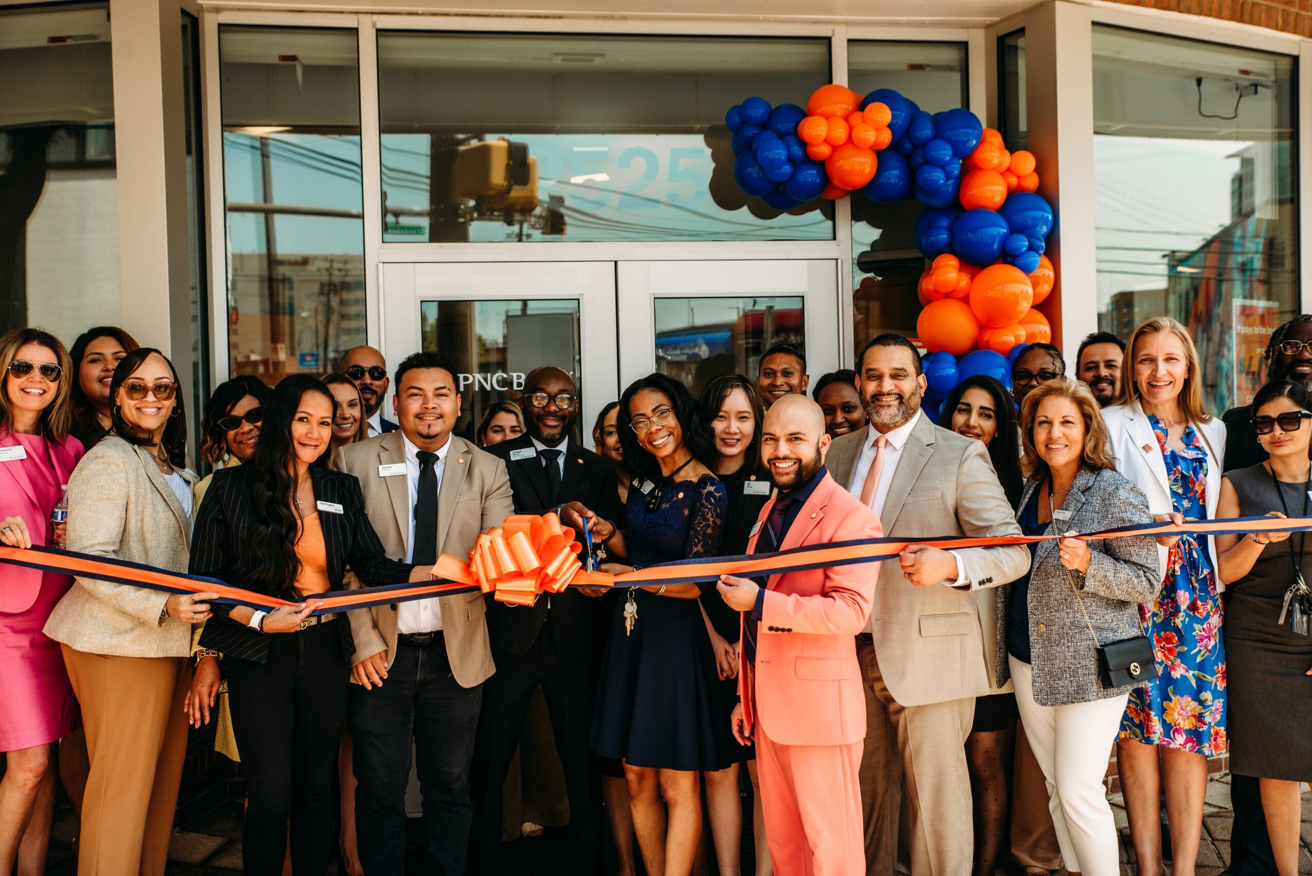 Group of diverse people celebrating outside a building with balloon decorations, holding a ribbon for a ribbon-cutting event.