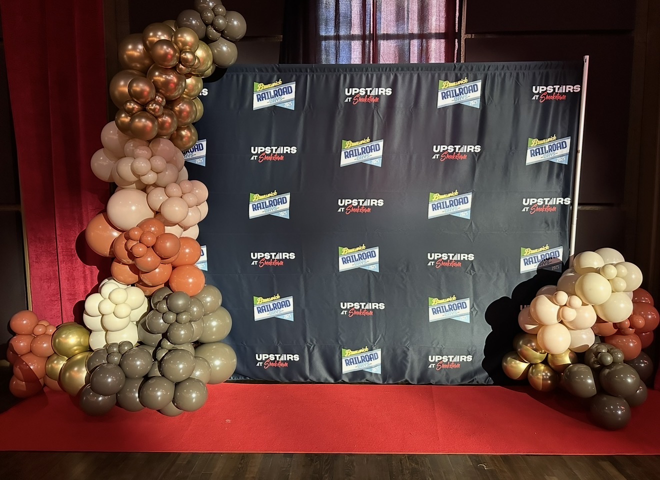 Balloon decorations on both sides of a black backdrop with logos, on a red carpeted floor.