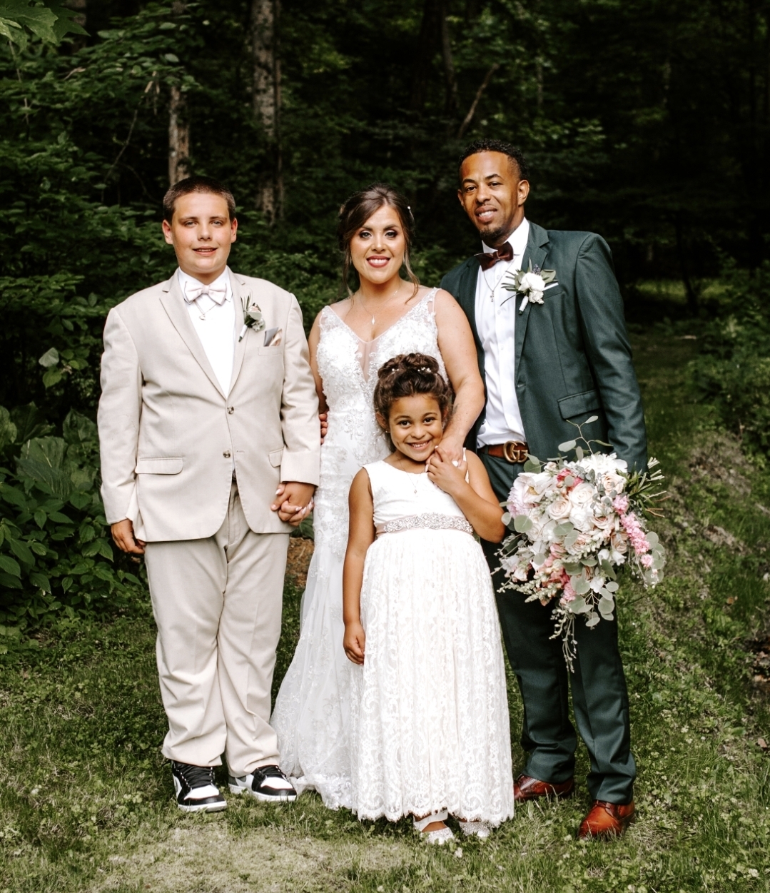 Four people in wedding attire standing outdoors in a forested area, smiling, with a woman holding a large bouquet of flowers.