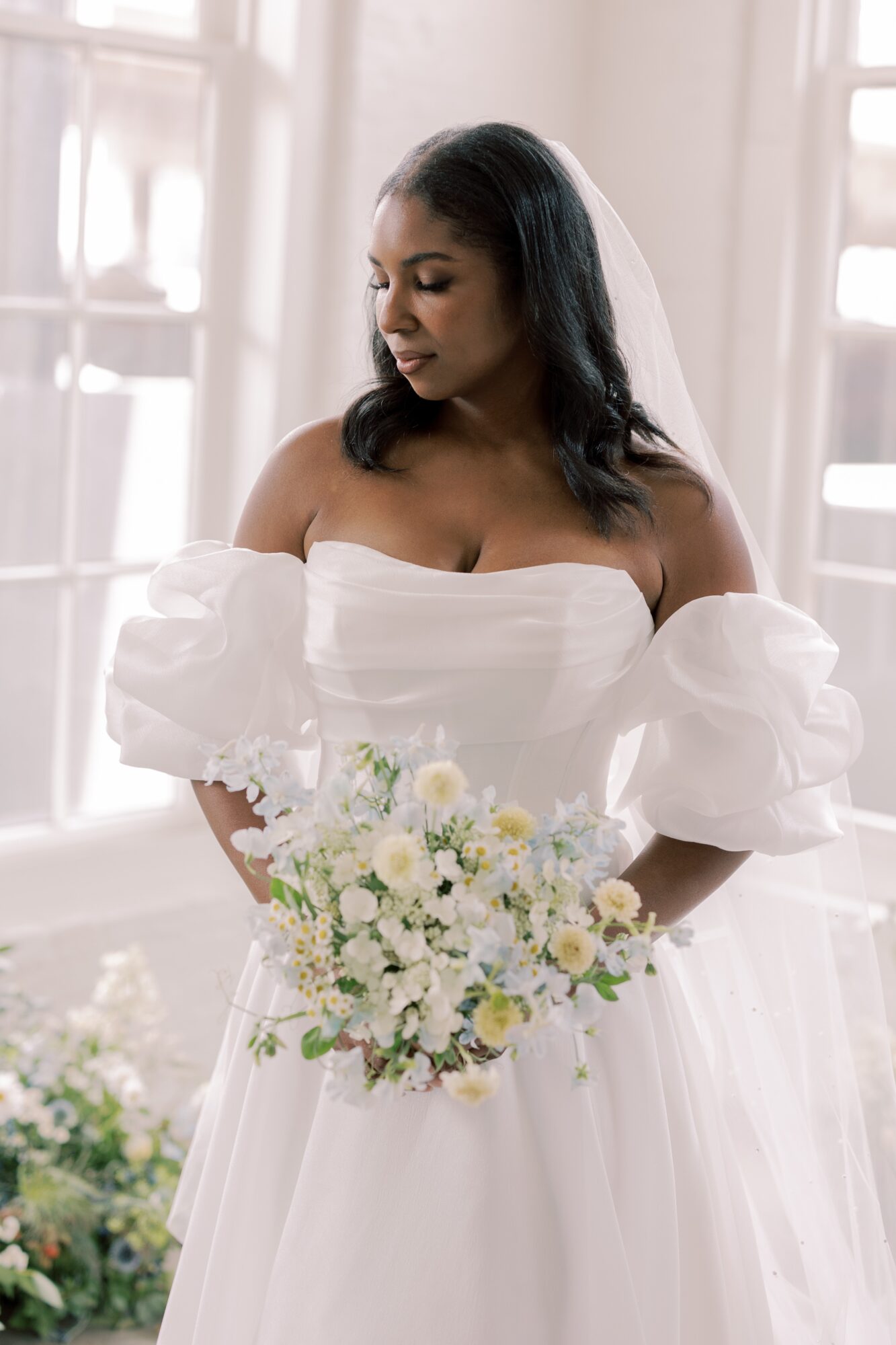 Woman in white dress holding a bouquet of white and yellow flowers, standing indoors near windows, looking down.