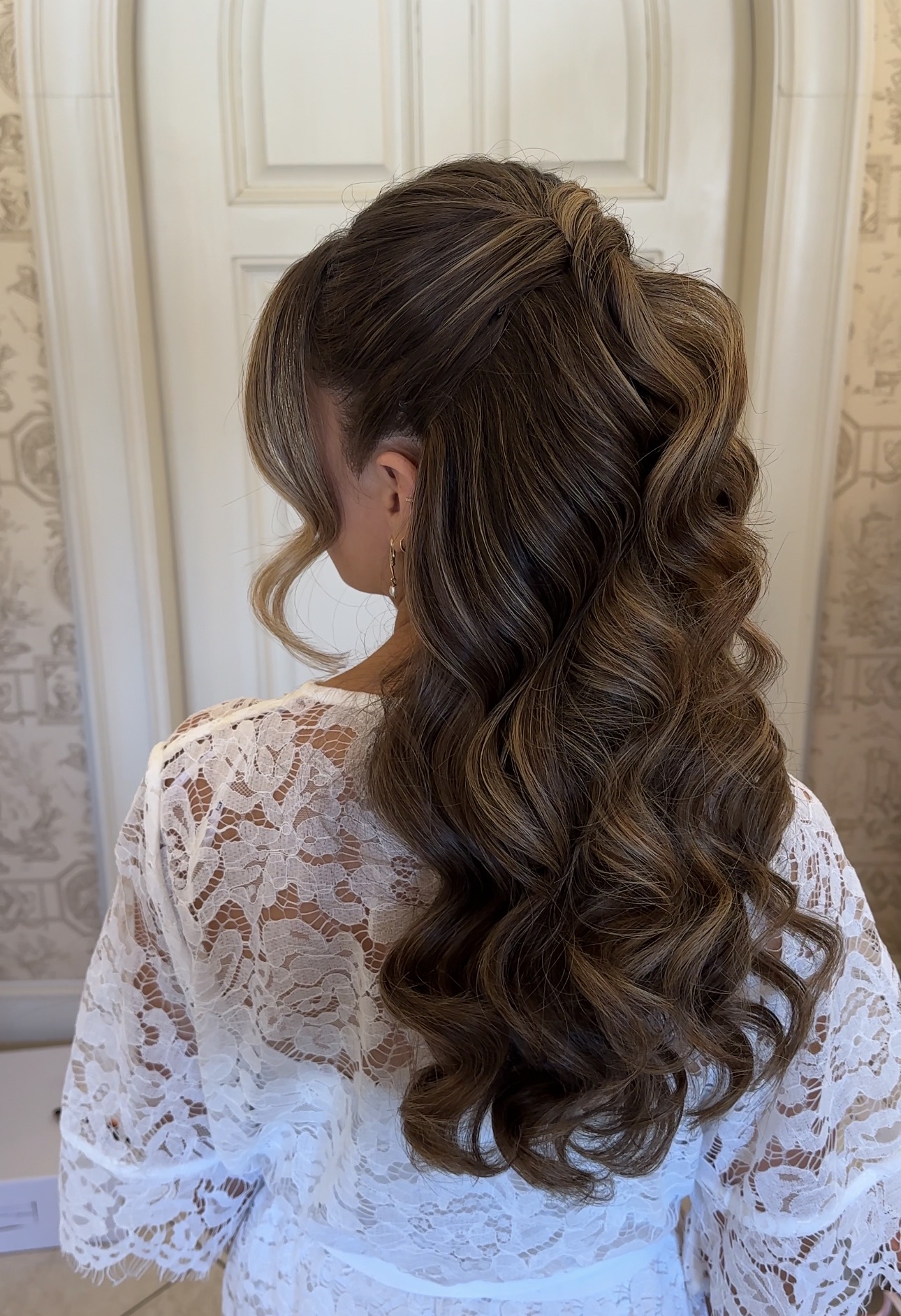 Woman with long, wavy brown hair styled in loose curls, wearing a white lace top, facing away from the camera.