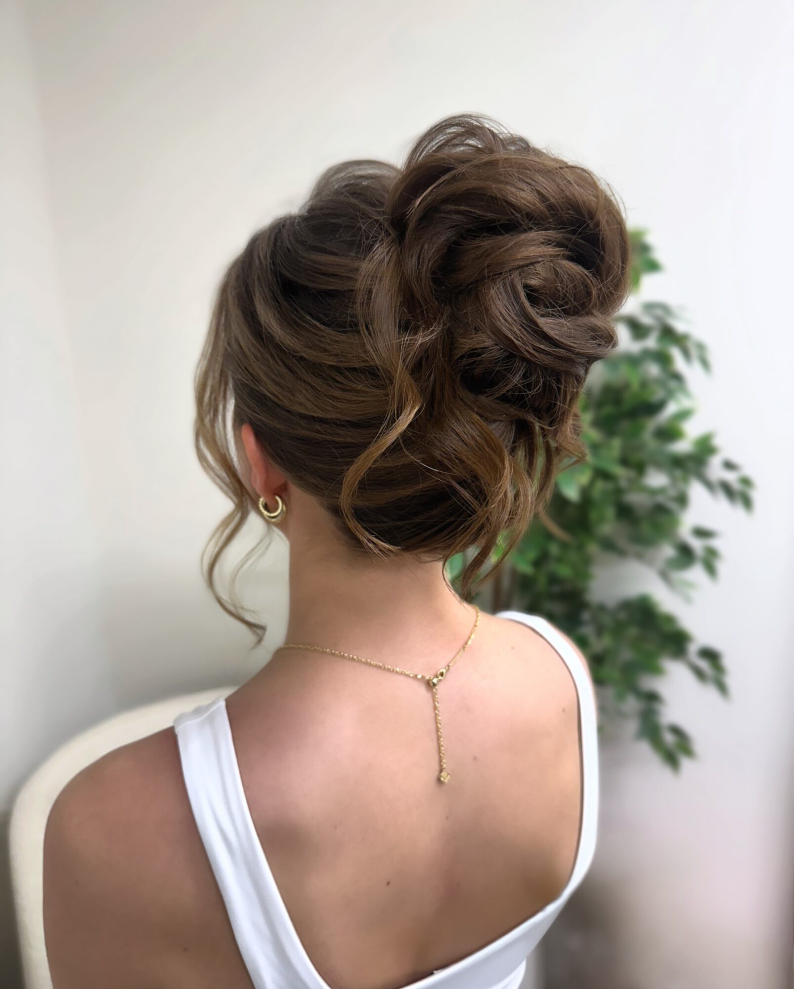 Back view of a woman with an elegant updo hairstyle, wearing a white top and gold jewelry, with a plant in the background.