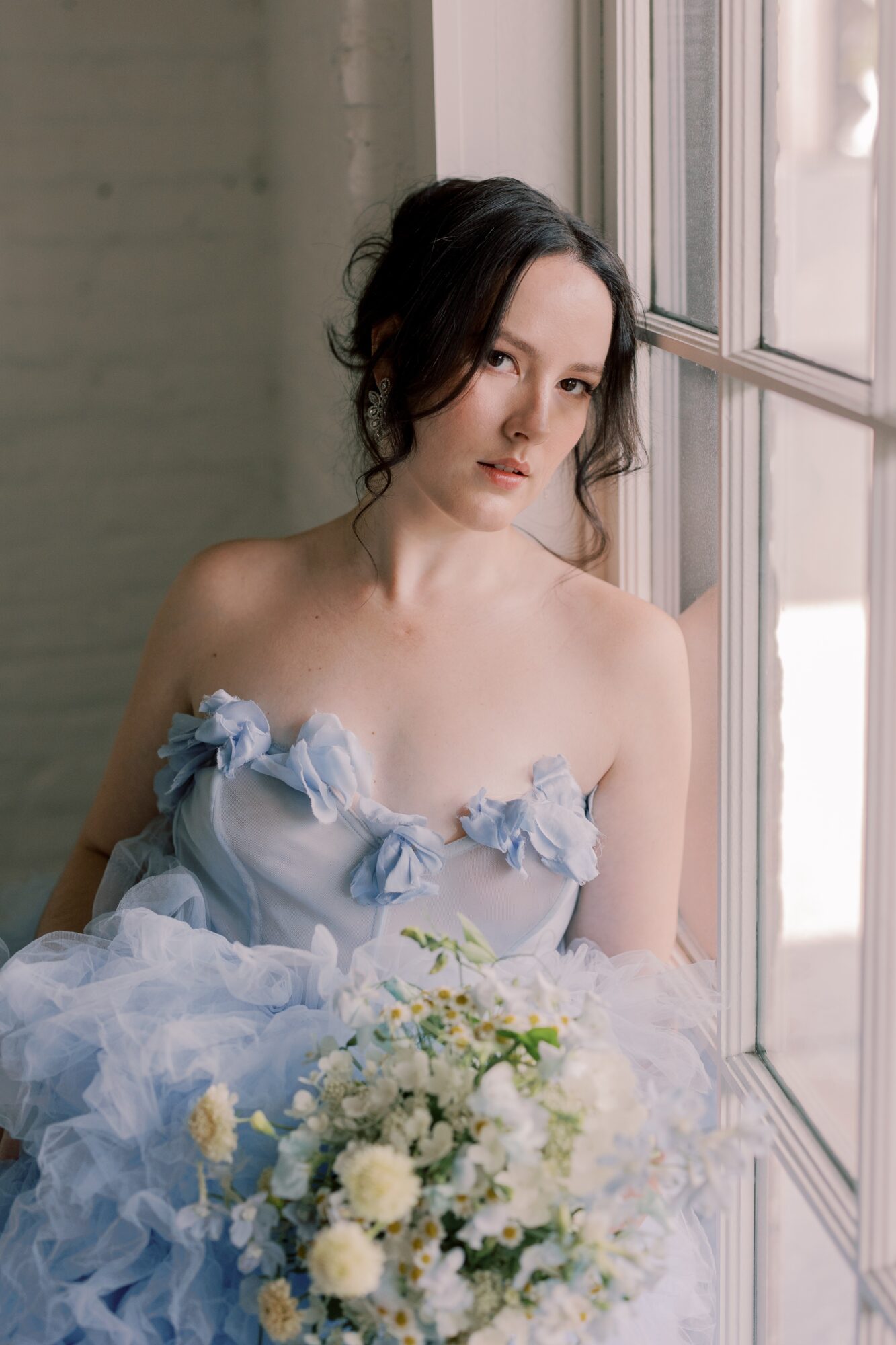 Young woman with dark hair in a loose updo, wearing a light blue dress with ruffles, holding a bouquet of white flowers, looking out a window.