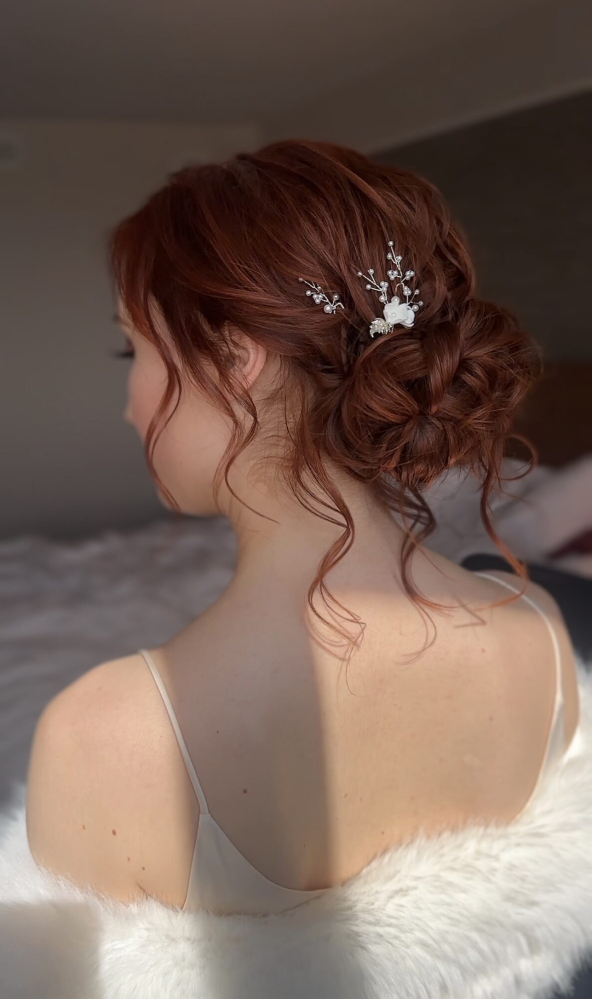 Side view of a woman with red hair styled in an updo with a decorative hairpiece, wearing a white dress.