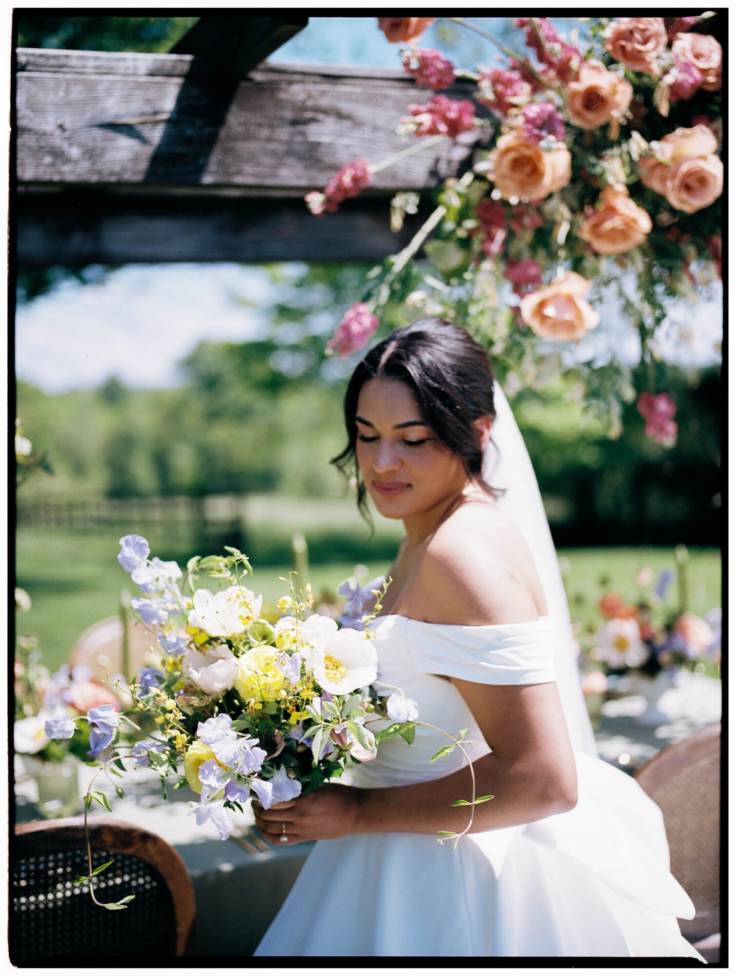 Young woman in white dress holding a bouquet of flowers outdoors, with a large floral arrangement overhead.