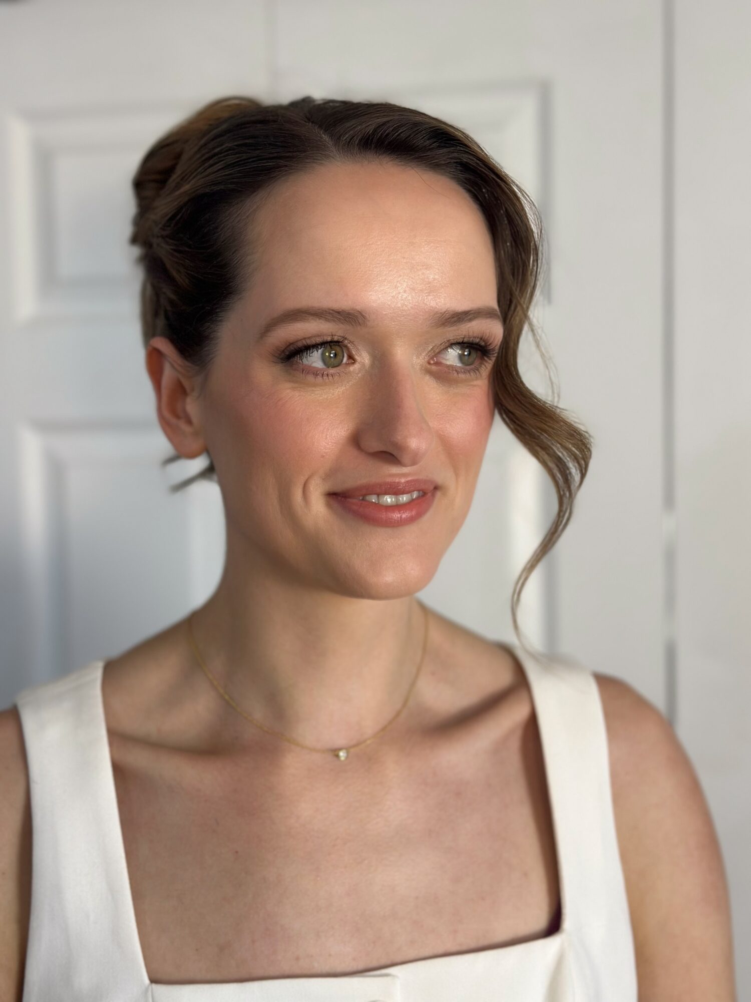 Young woman with styled hair and makeup, wearing a white sleeveless top, smiling softly, standing indoors near a door.