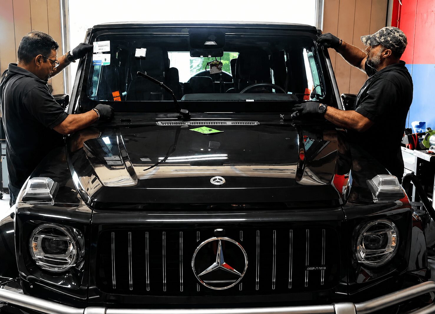 Two men work on a black Mercedes-Benz vehicle inside a workshop, handling the windshield. The vehicle's front grille and logo are visible.