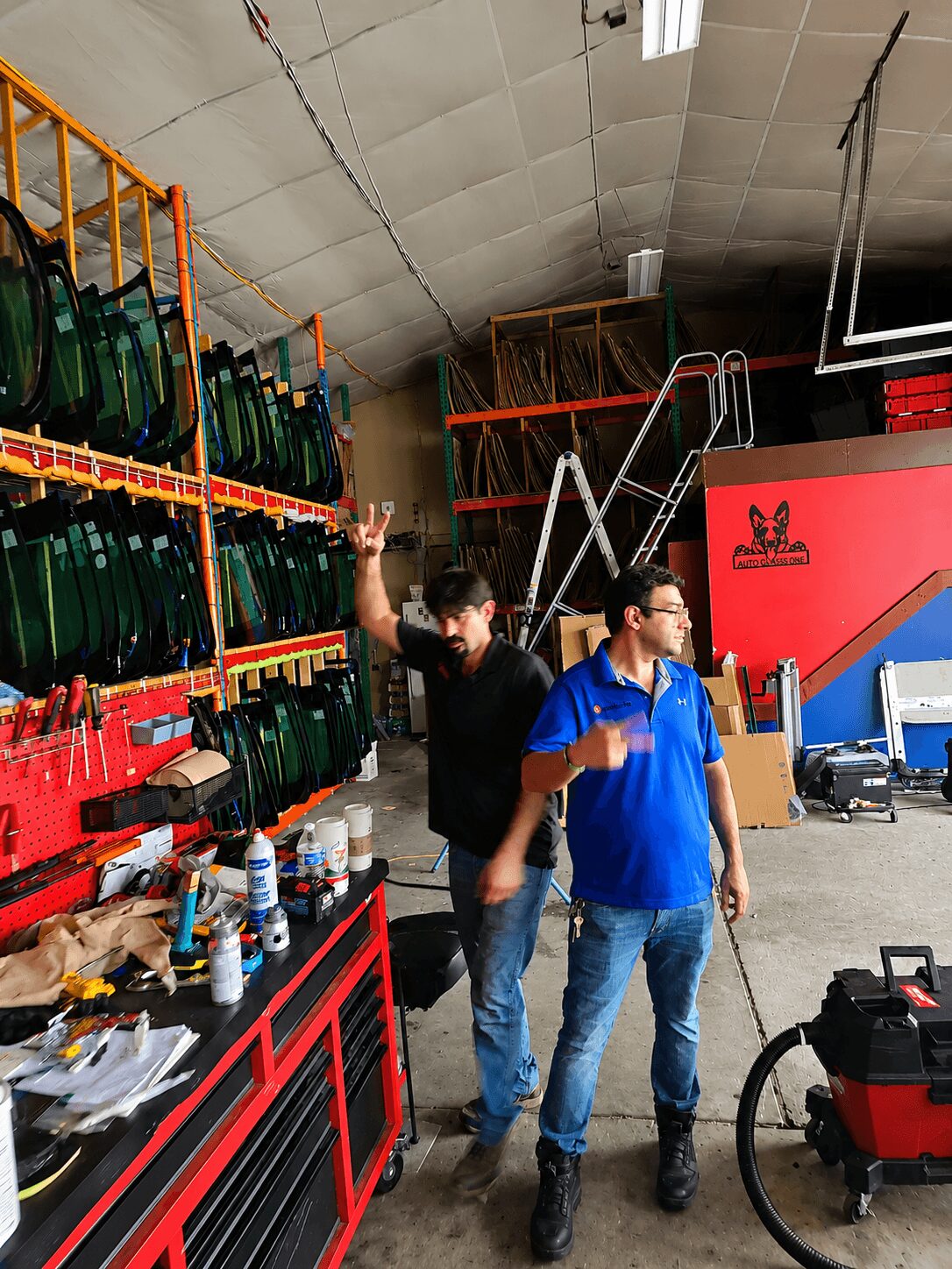 Two men in a warehouse with shelves of glass bottles, one making a peace sign, the other looking away, industrial setting.