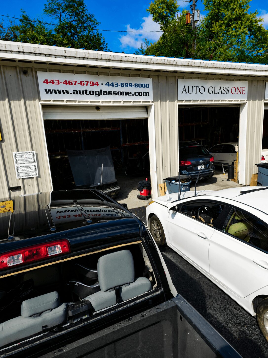 Auto glass shop with open garage doors, cars parked outside, and a sign displaying contact information and website.